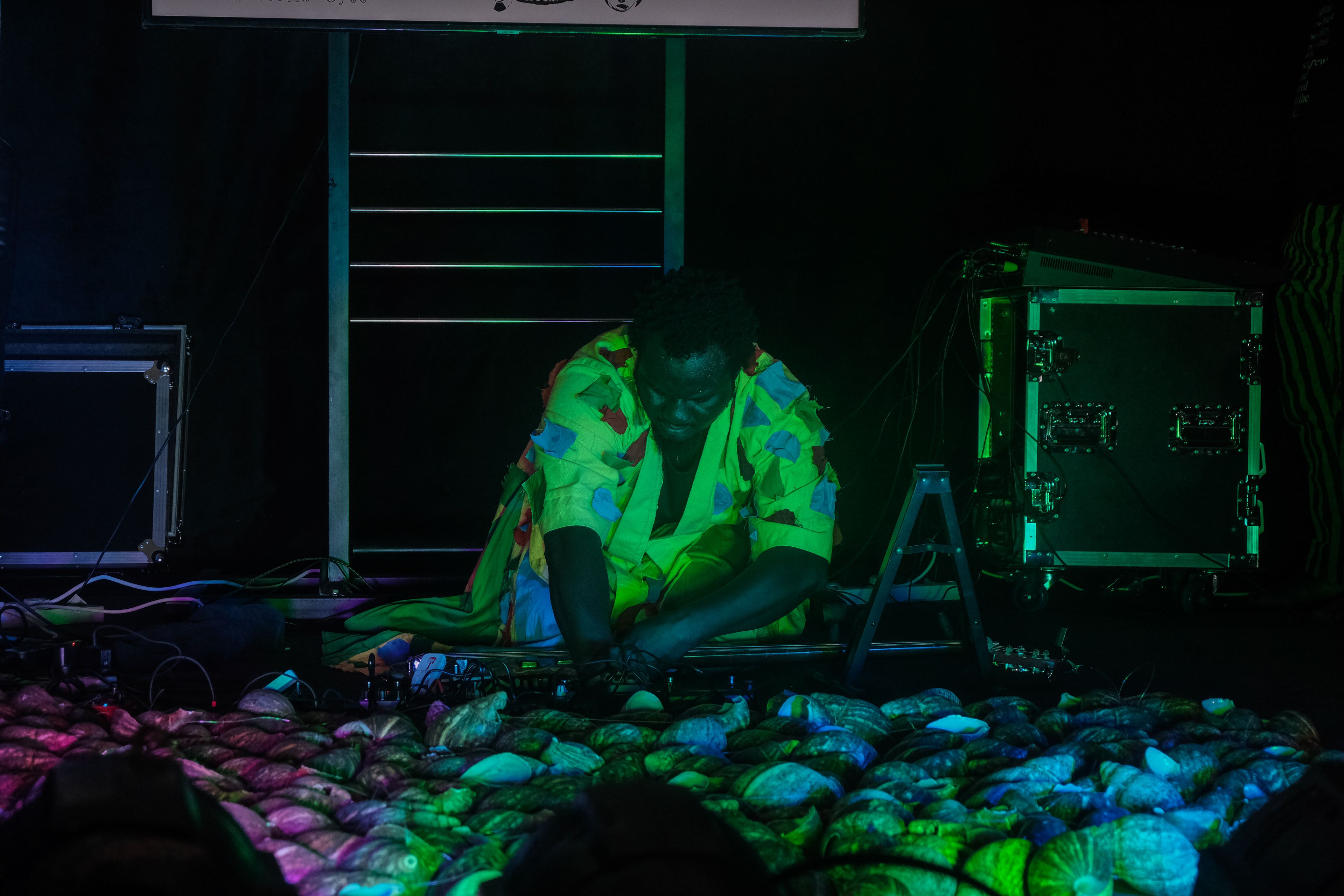 A person in a bright green patterned outfit kneels on a dark stage, interacting with a sprawling arrangement of shells illuminated by purple and green light.