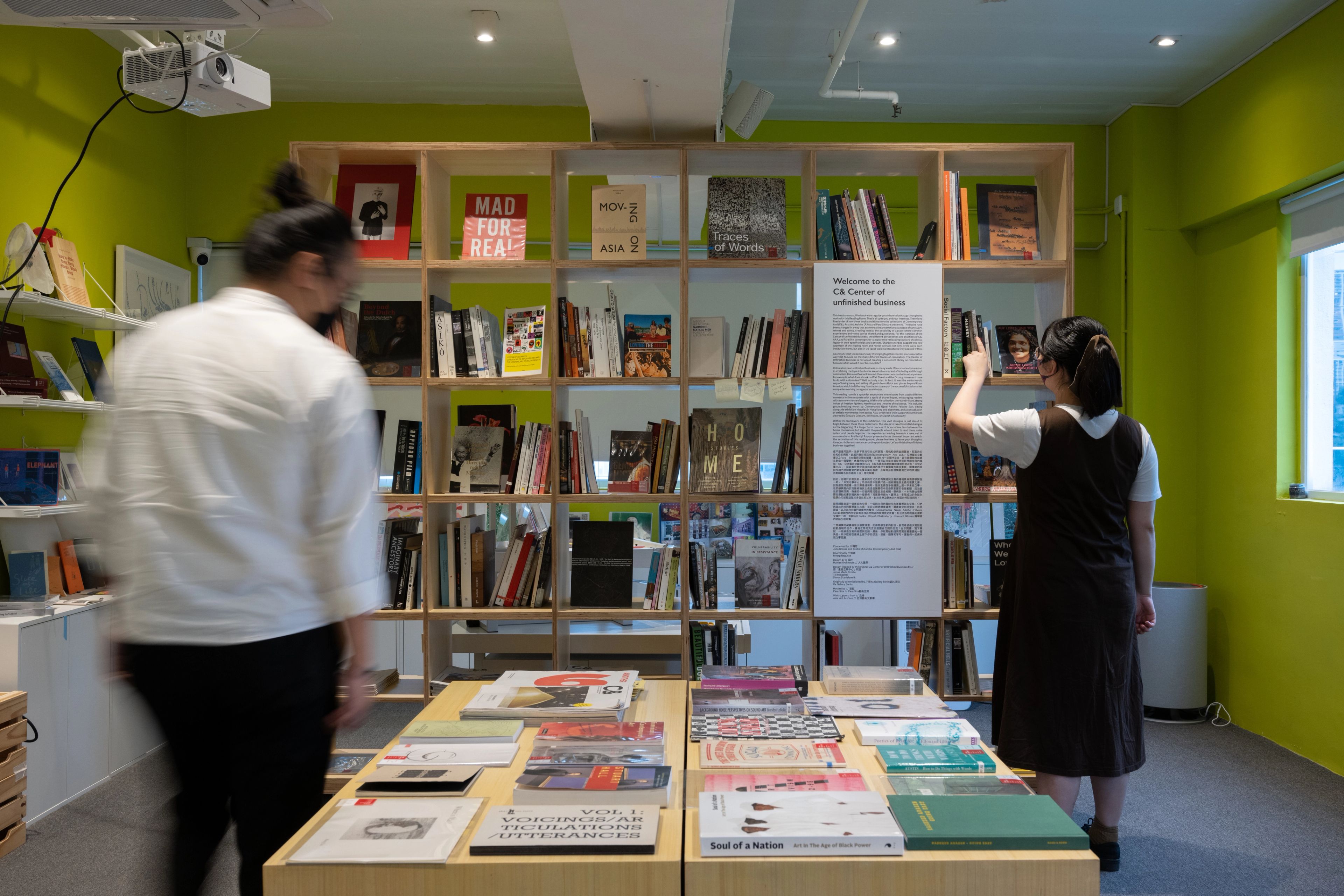 Two people in a bright green room, one blurred walking past a table of books, the other browsing a large bookshelf filled with books.