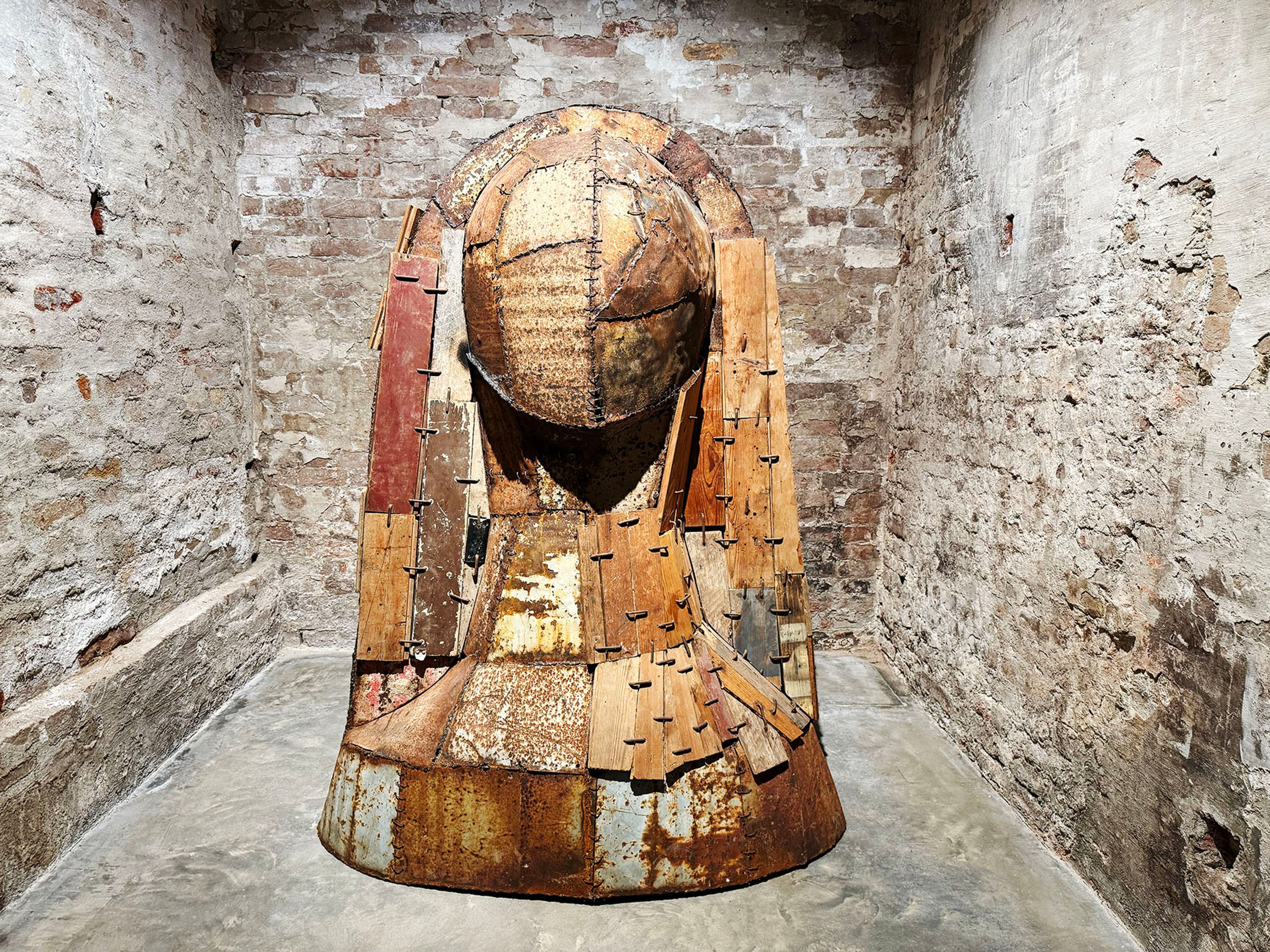 A towering, weathered sculpture made of salvaged wood planks and rusted metal sheets, resembling a hooded figure, stands in a room with rough brick walls.