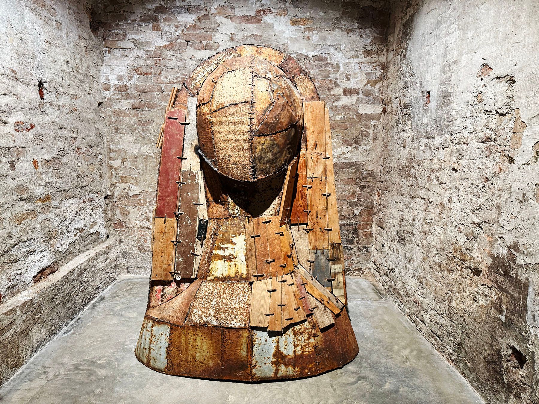 A towering, weathered sculpture made of salvaged wood planks and rusted metal sheets, resembling a hooded figure, stands in a room with rough brick walls.