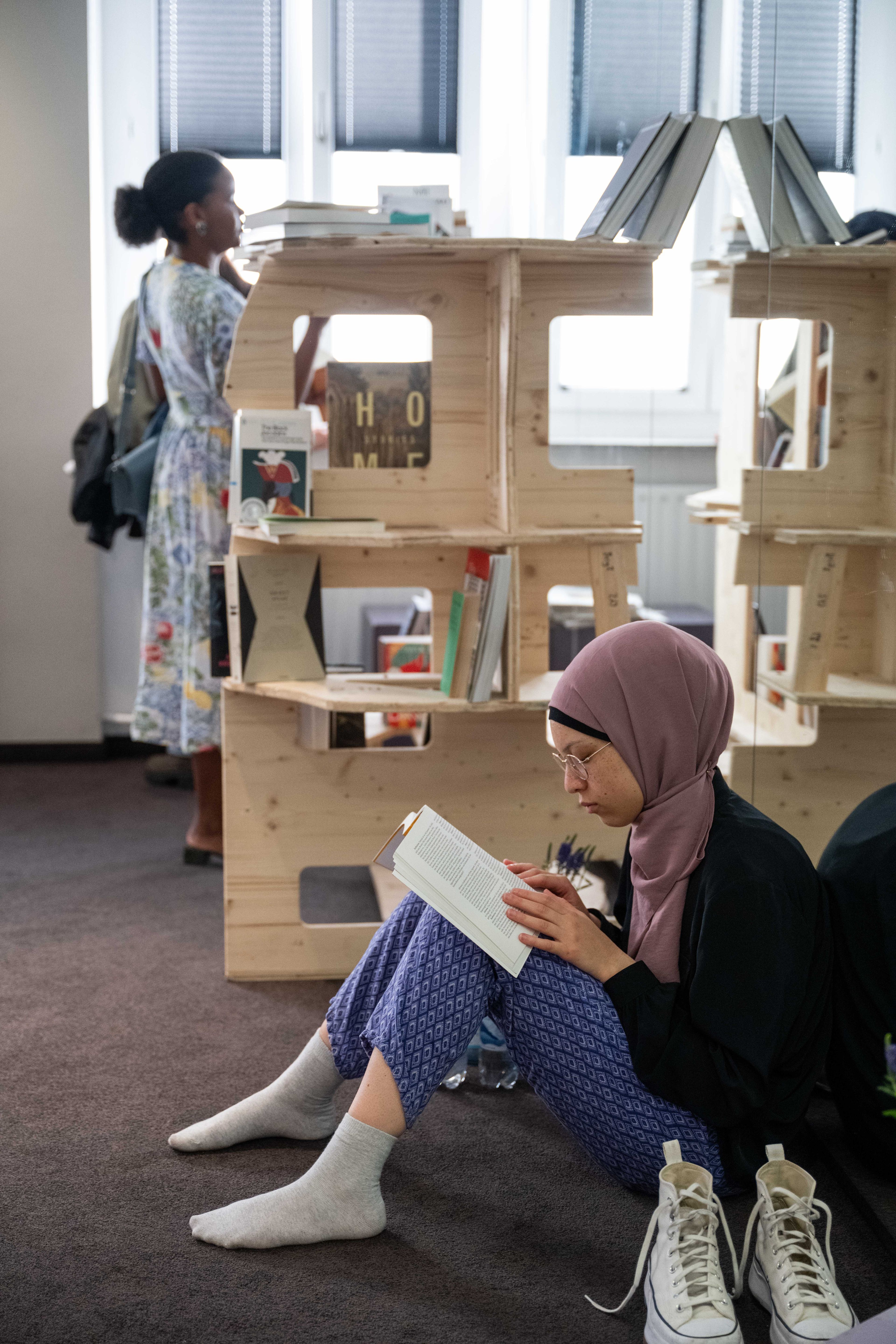 A woman in a pink hijab sits on the floor reading a book, with wooden modular bookshelves and another person in the background.