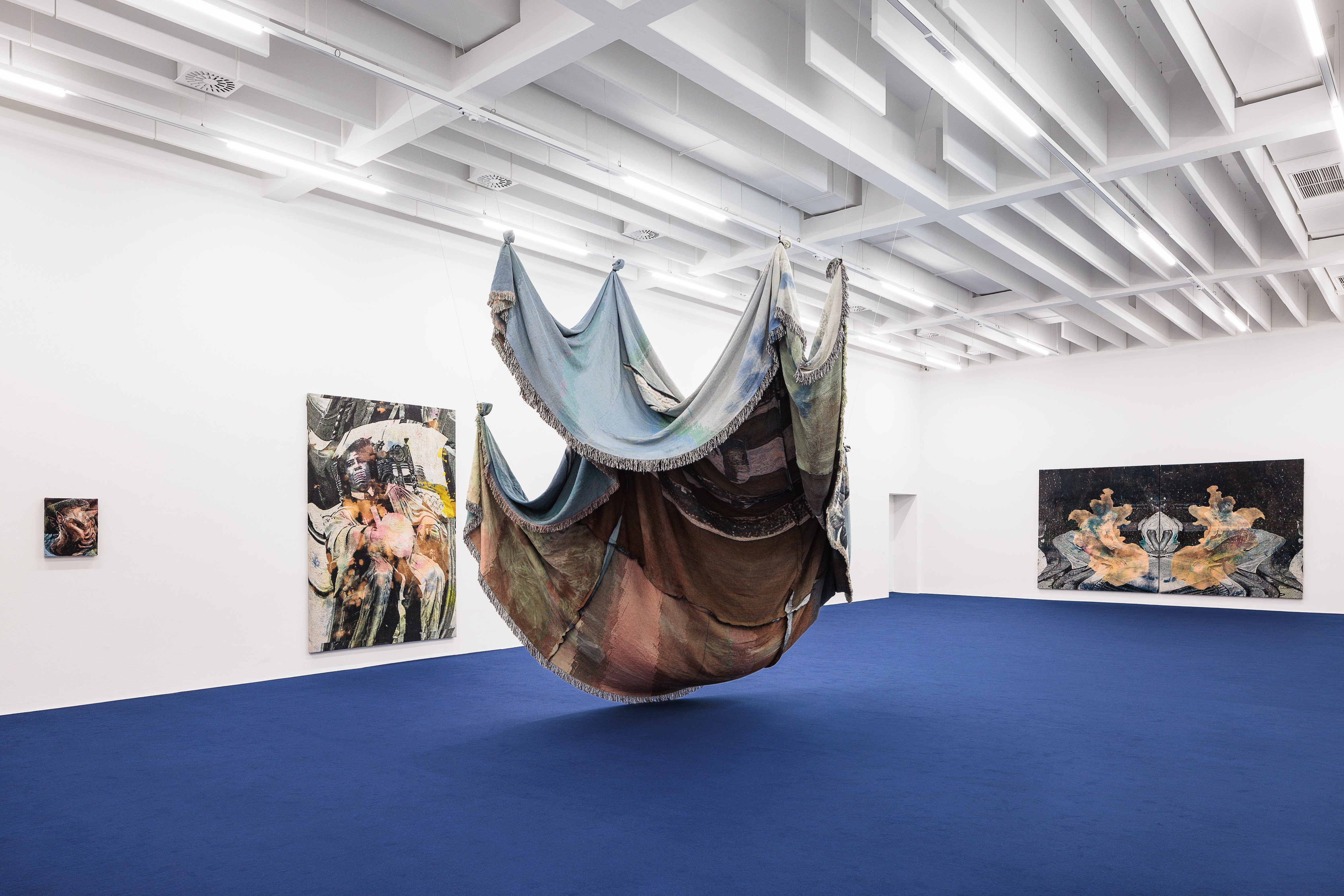 A modern art gallery with a blue carpet, featuring a large, draped textile sculpture suspended from the ceiling, surrounded by paintings on white walls.