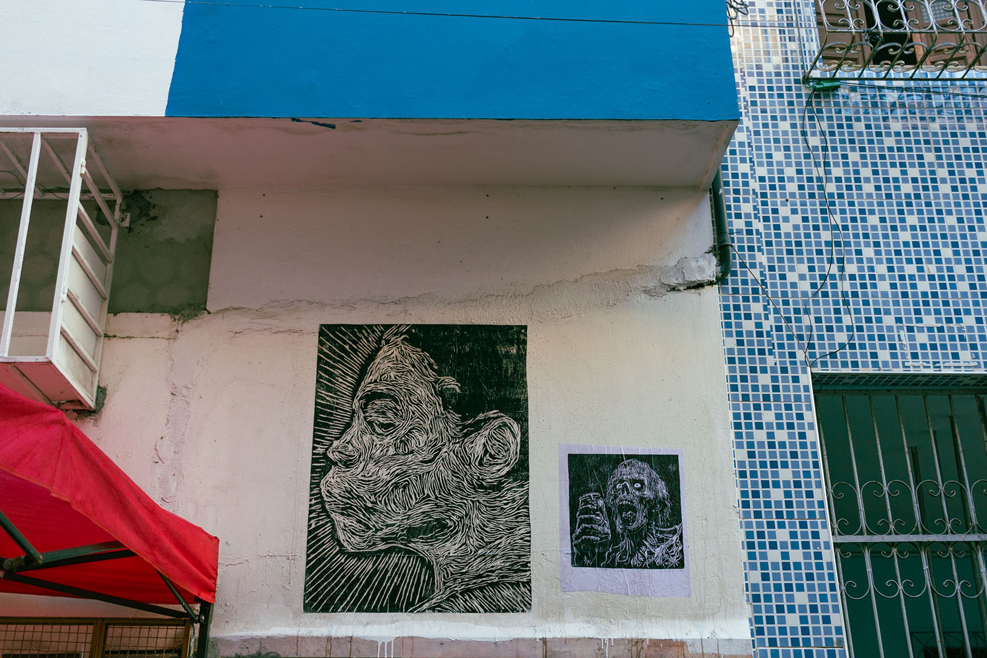 Two black and white linocut-style artworks of expressive faces on a light-colored building wall, beneath a blue ledge and next to a blue tiled section.