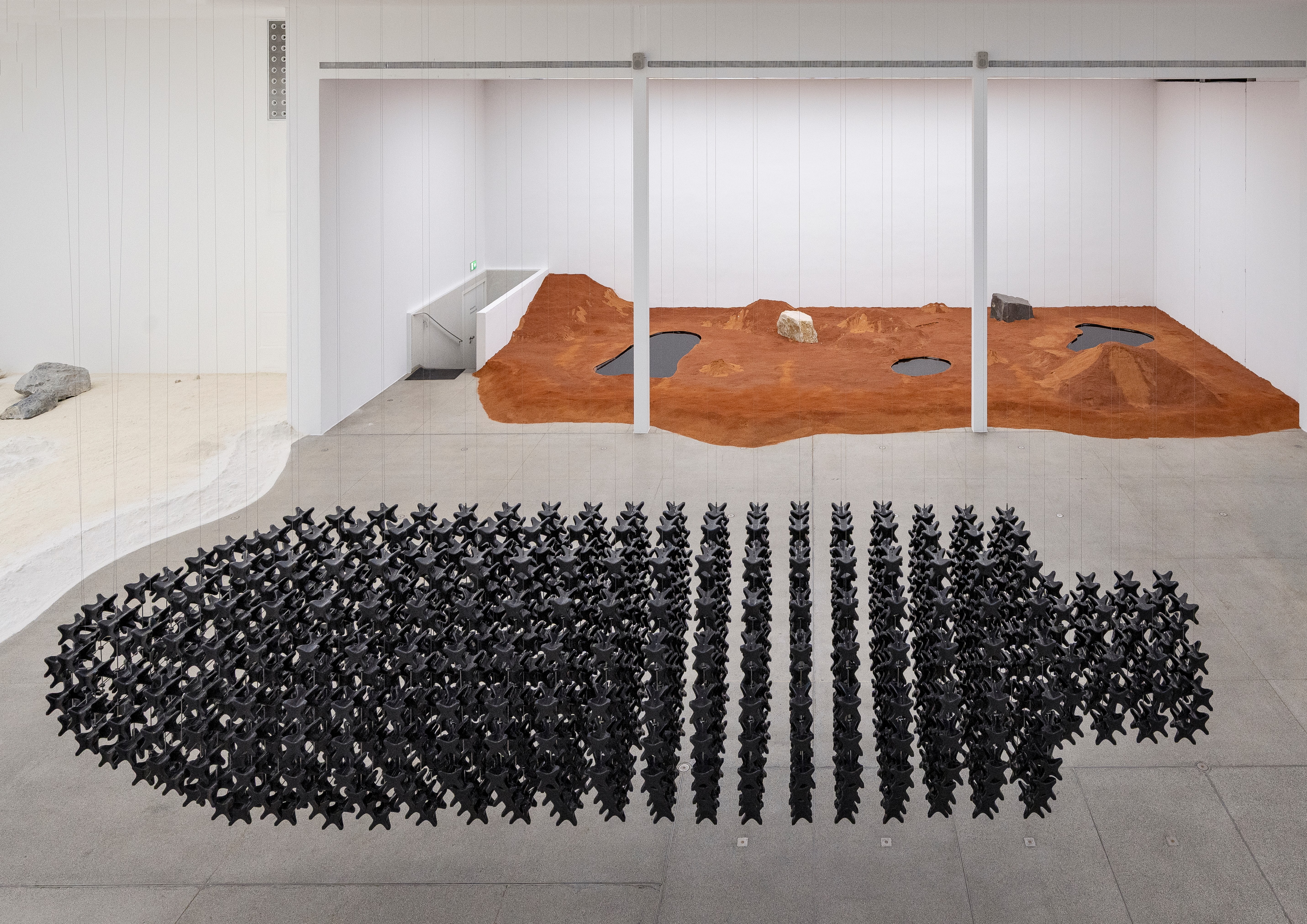 Large, dark, spiky, torpedo-shaped sculpture on a gray floor. Behind it, a raised reddish-brown landscape sculpture features dark pools and rocks within a white gallery.