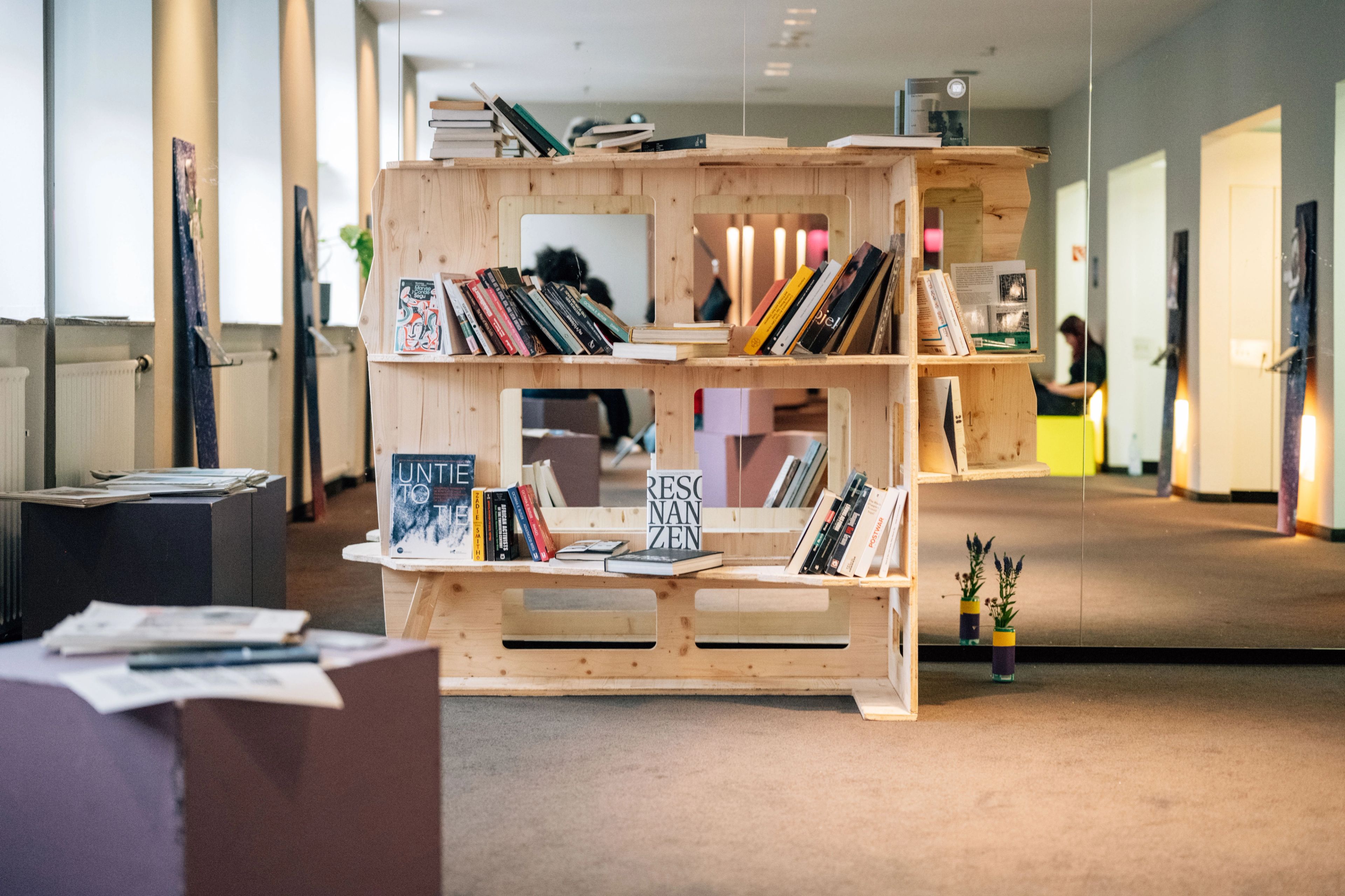 A light wooden bookshelf filled with books stands in a spacious room with a large mirror wall.