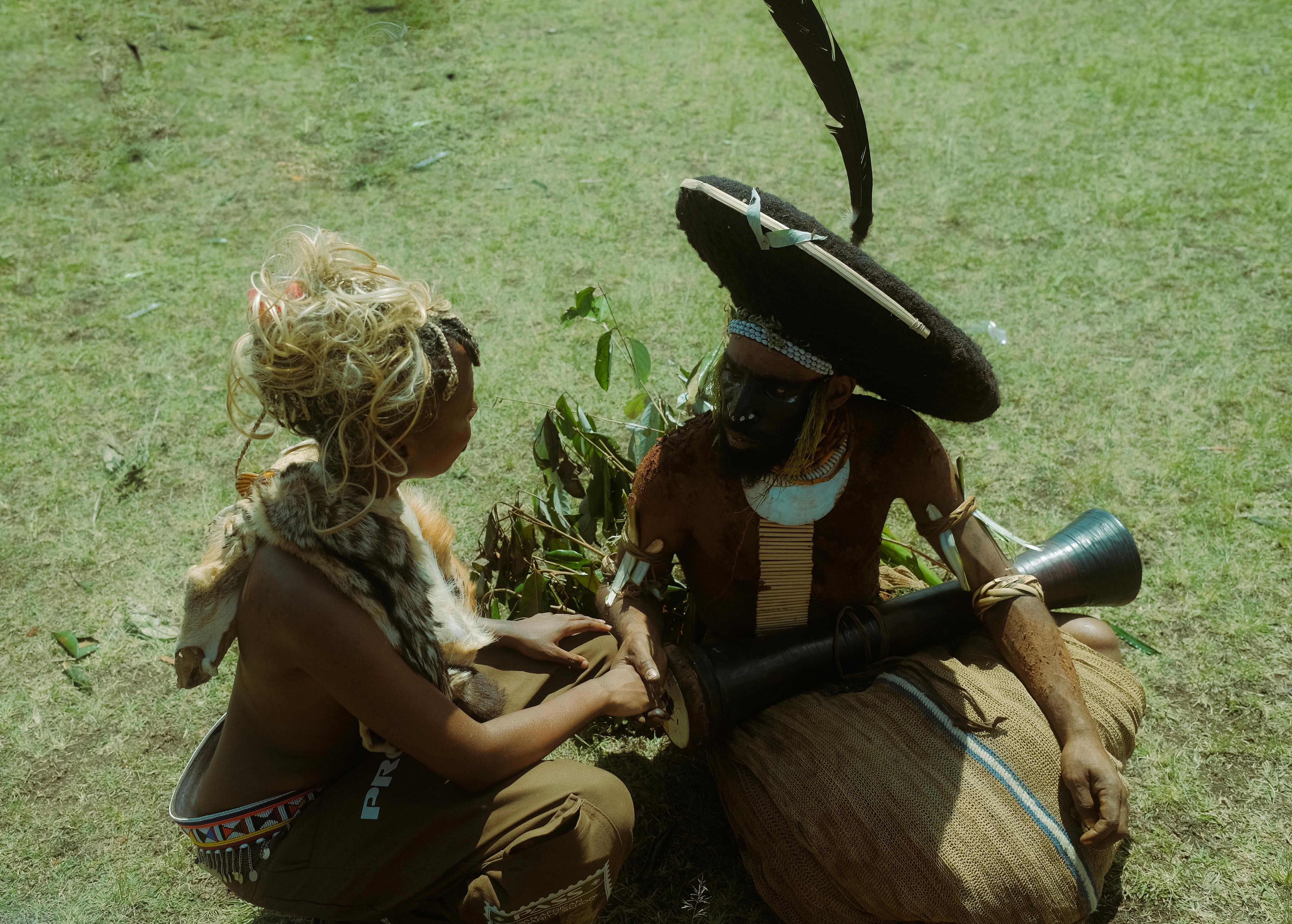 Two individuals in traditional attire on grass, one with a fur pelt, the other with face paint and a drum.
