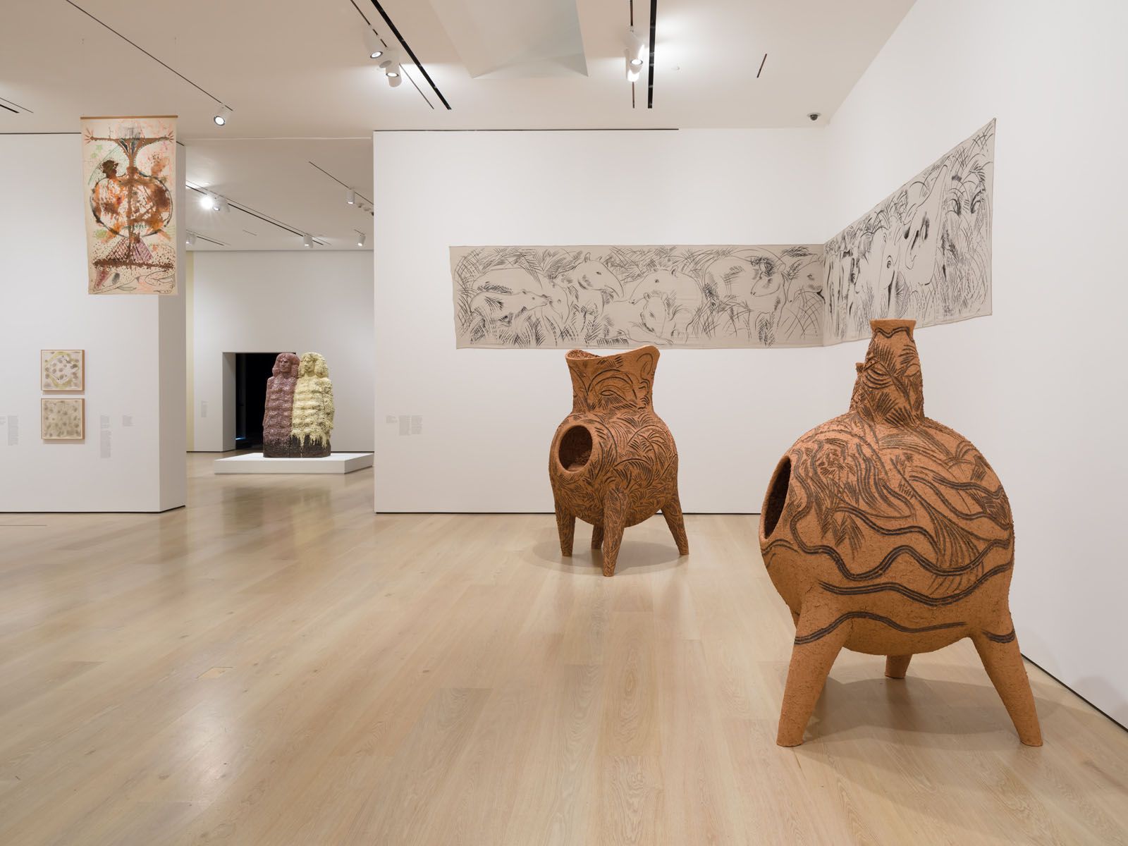 An art gallery featuring two large, patterned terracotta sculptures in the foreground, a panoramic wall drawing, and other diverse artworks.