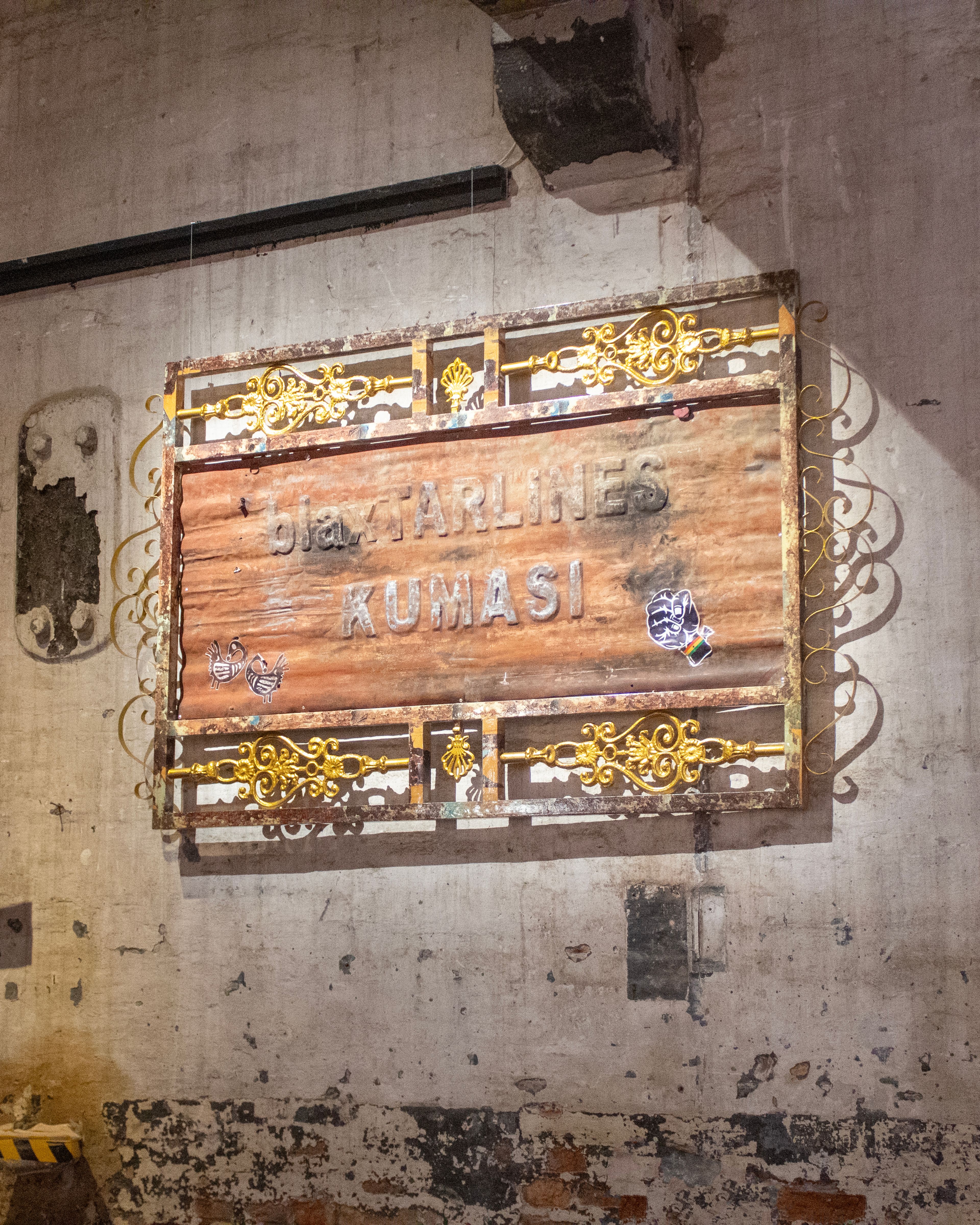 Distressed wooden sign with embossed text 'Bla GARLINES KUMASI' in an ornate, rusty gold metal frame on a textured wall.