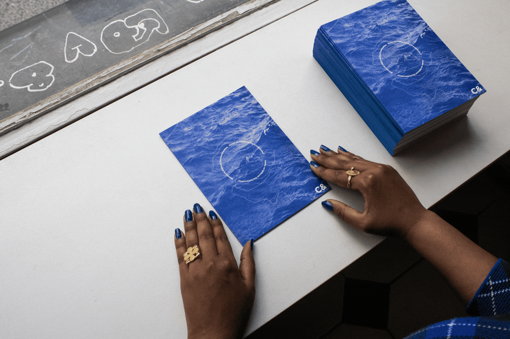 Hands with blue nail polish hold a blue publication with a wave pattern and a circle graphic, next to a stack of identical cards.