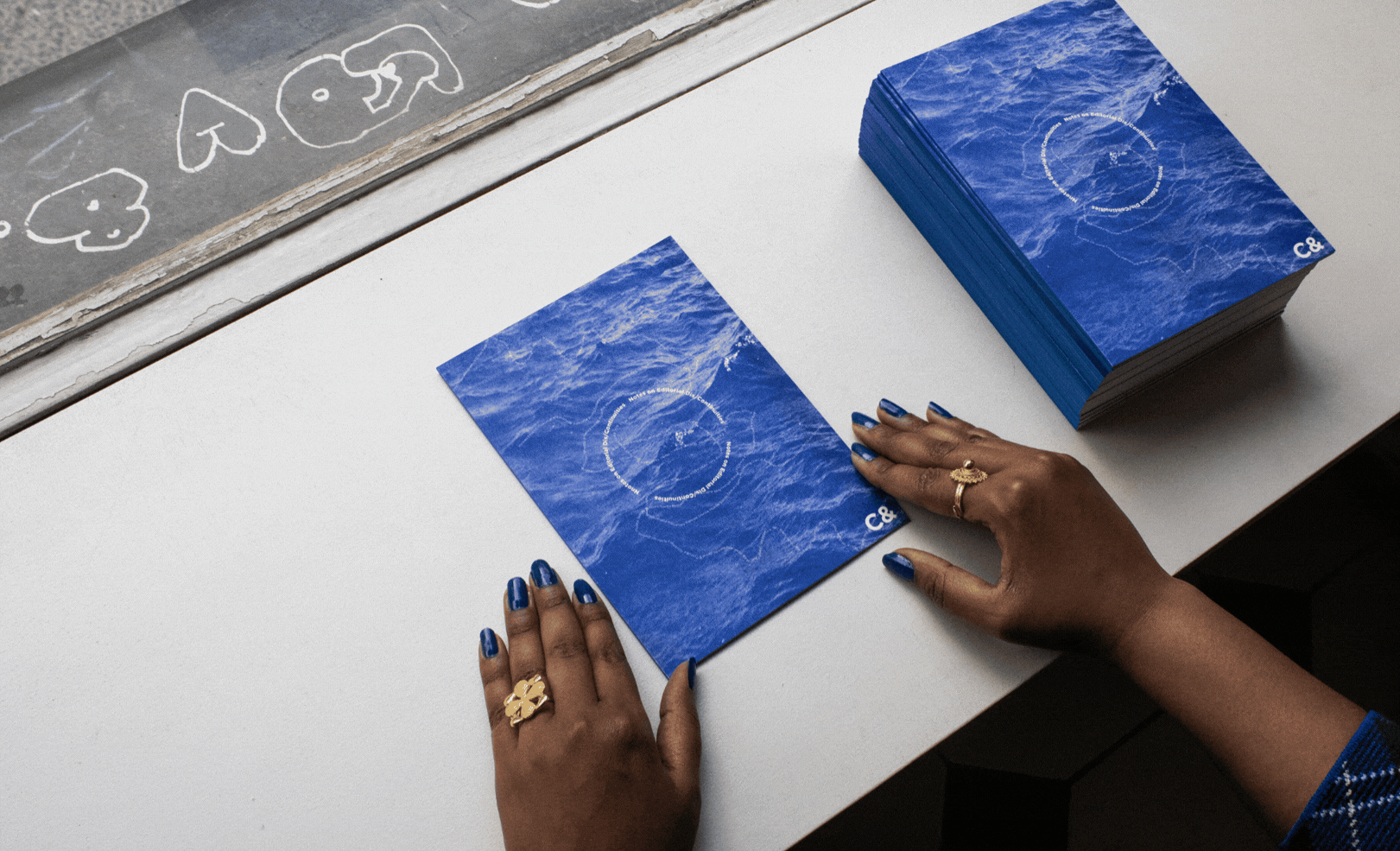 Hands with blue nail polish hold a blue publication with a wave pattern and a circle graphic, next to a stack of identical cards.