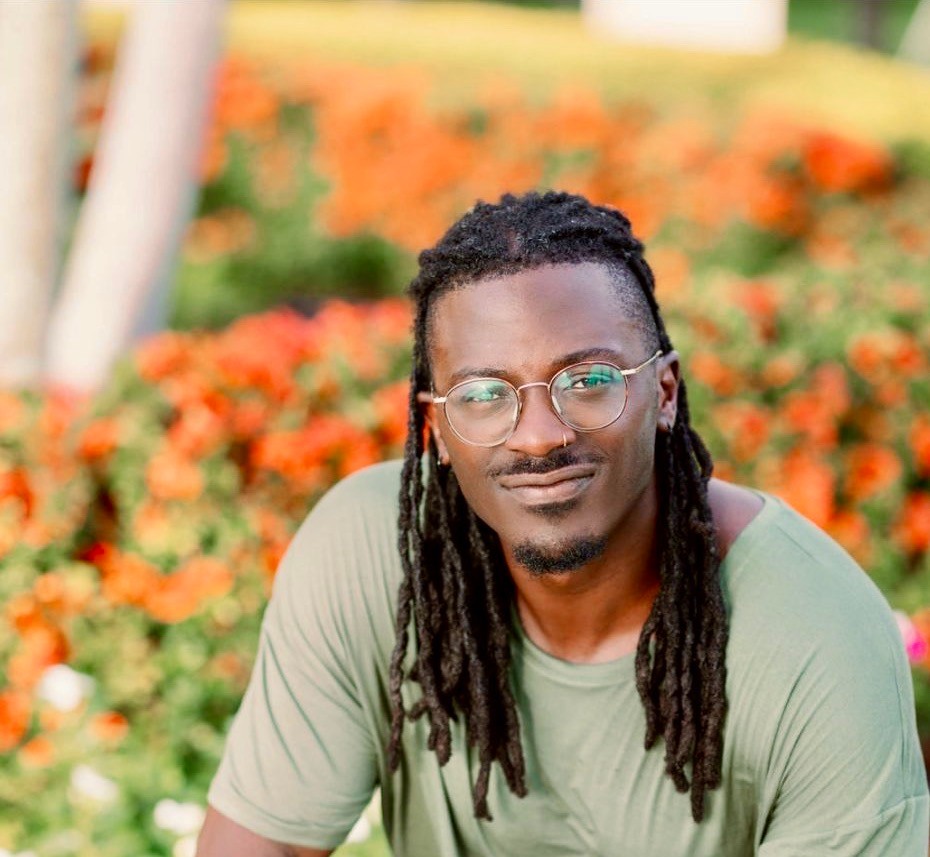 A Black man with dreadlocks and glasses smiles faintly against a blurred background of orange flowers.