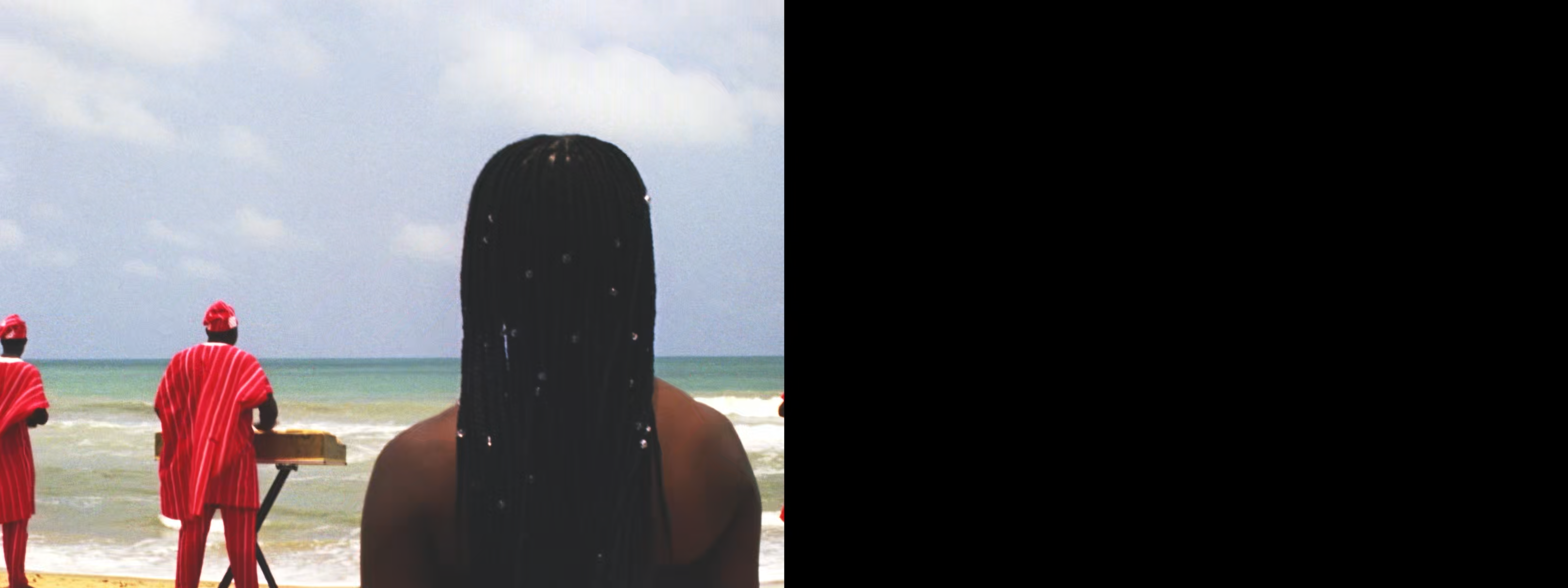 Rear view of a woman with long braids watching two people in red by the ocean. The right half of the image is black.