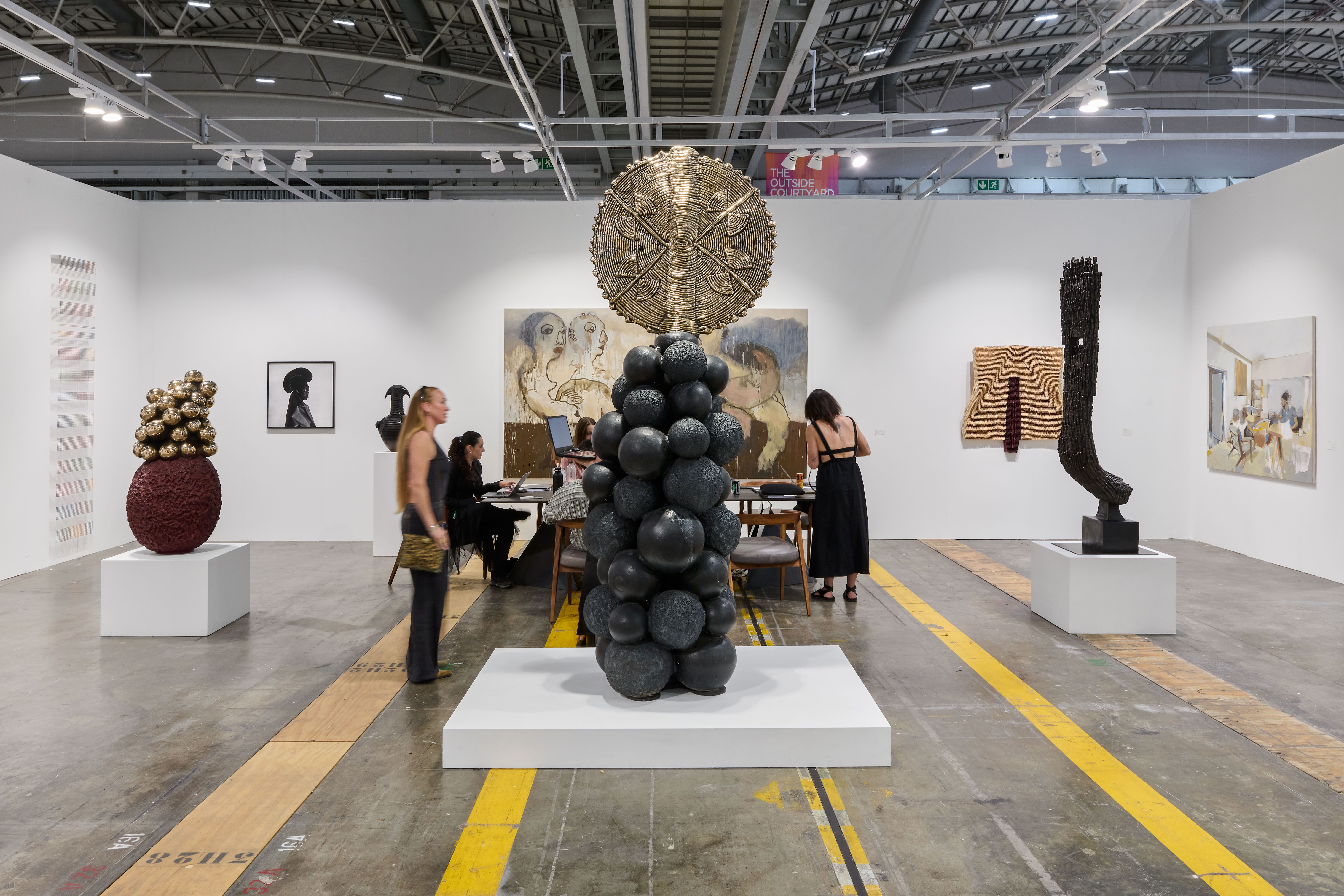 A spacious contemporary art gallery with multiple abstract sculptures, paintings on white walls, and people viewing the works.