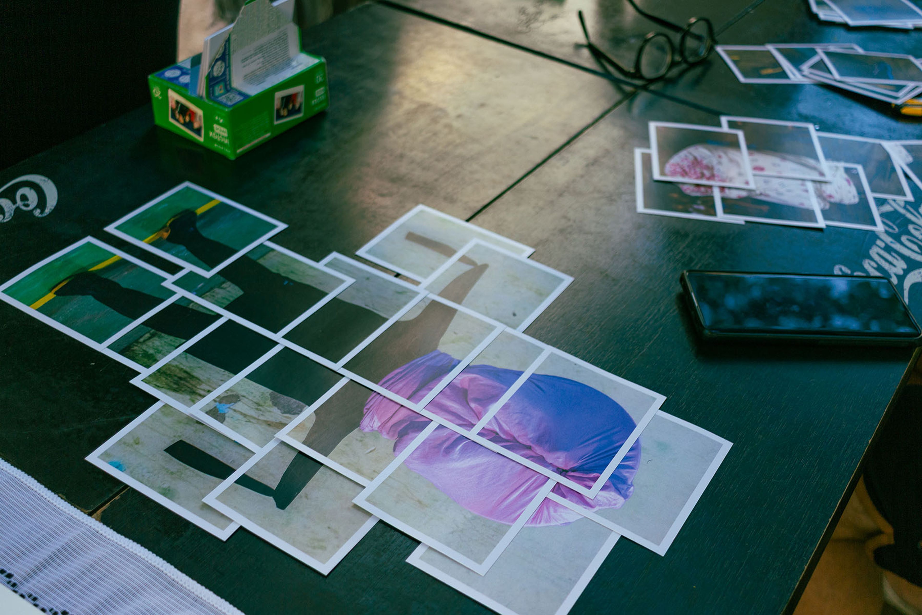 Small printed photos arranged on a dark table, some forming larger images, next to a smartphone and glasses.