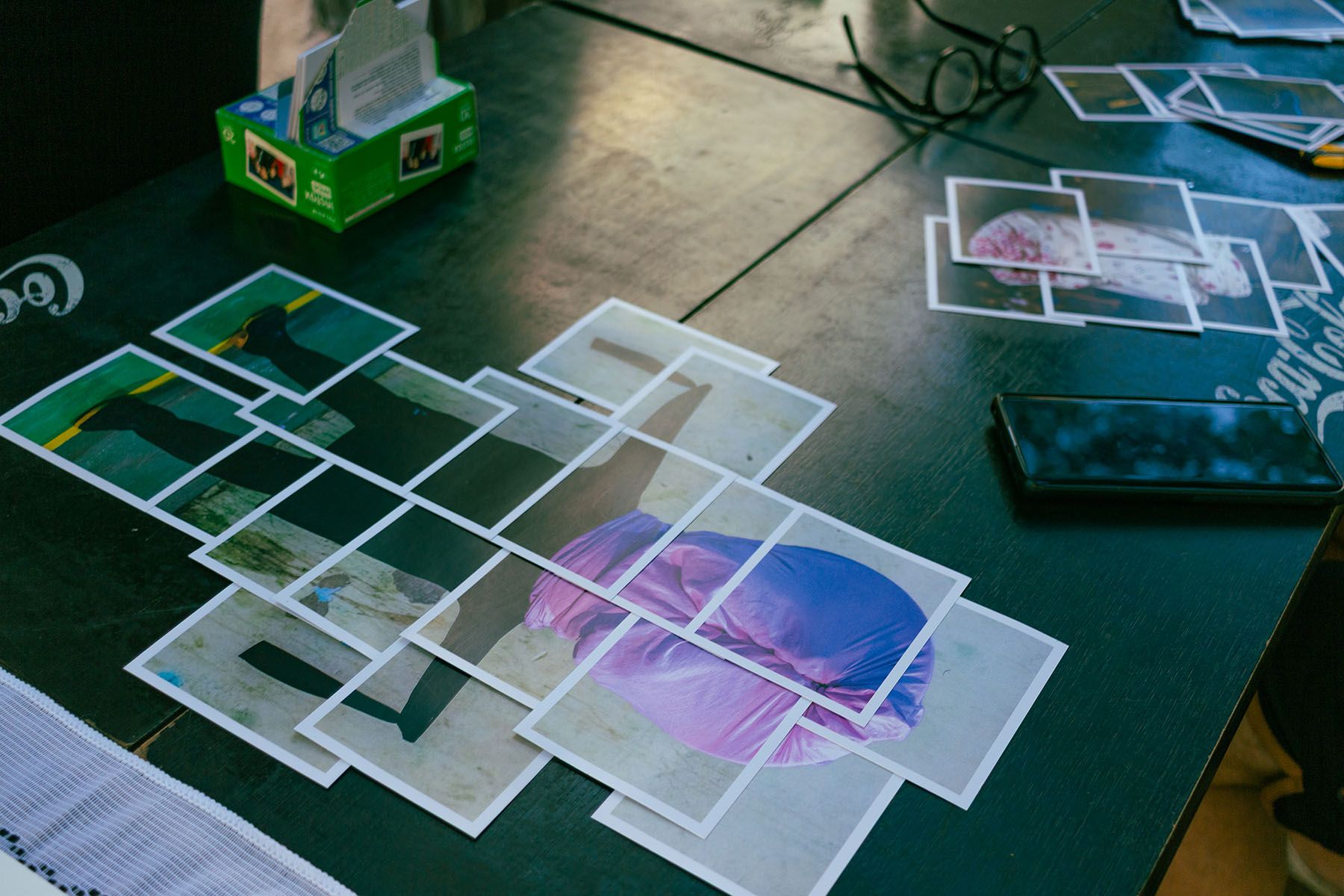 Small printed photos arranged on a dark table, some forming larger images, next to a smartphone and glasses.