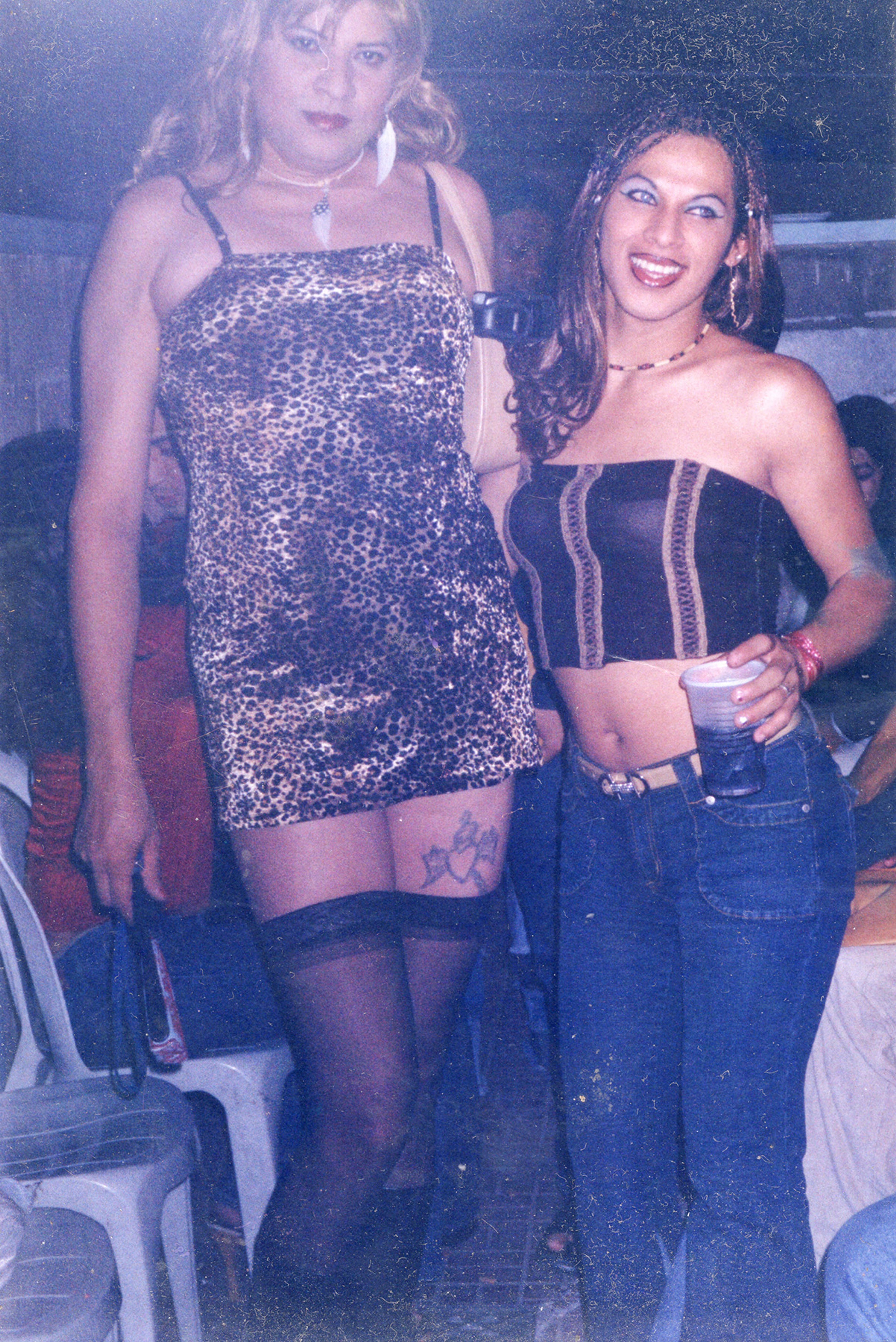 Two women at a party. The woman on the left wears a leopard print dress and black stockings. The woman on the right smiles in a strapless black crop top, blue jeans, and holds a drink.