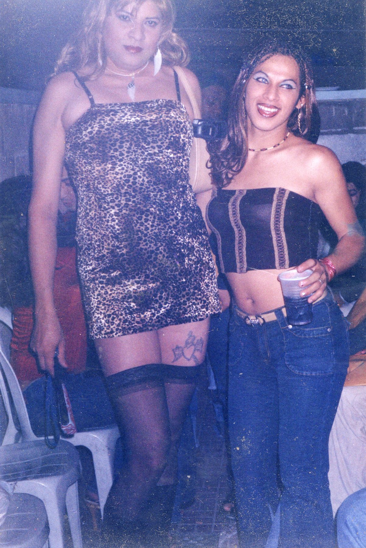 Two women at a party. The woman on the left wears a leopard print dress and black stockings. The woman on the right smiles in a strapless black crop top, blue jeans, and holds a drink.