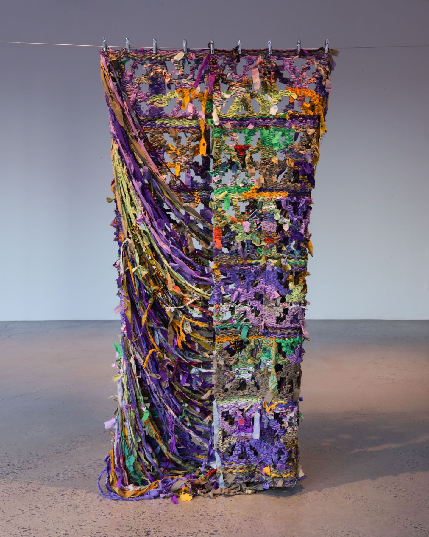 A tall, vibrant textile sculpture of woven purple, green, and orange fabric strips, hanging from a rod.