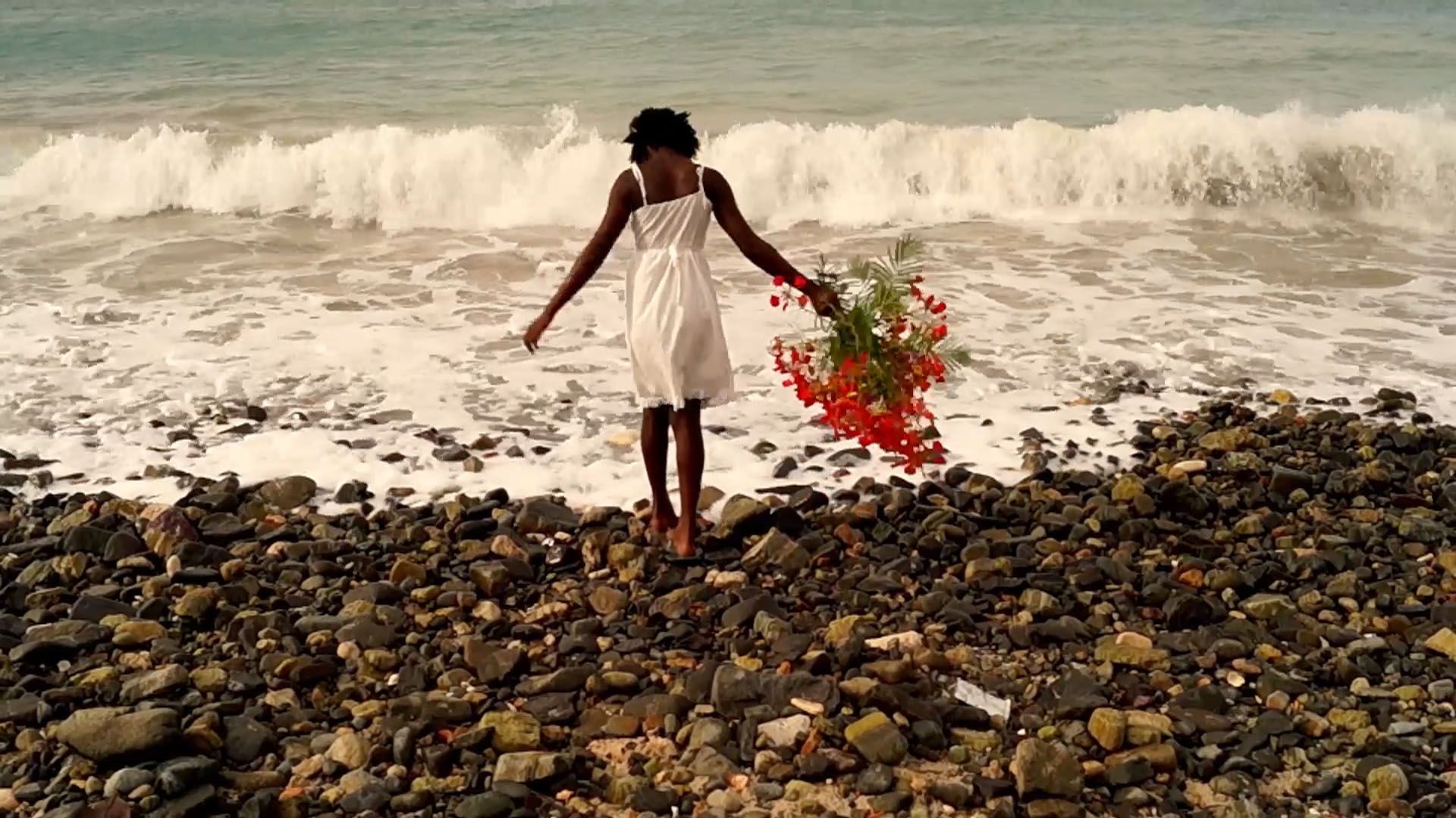 A person in a white dress walks barefoot on a rocky beach, carrying a large bouquet of red flowers, with ocean waves crashing behind them.