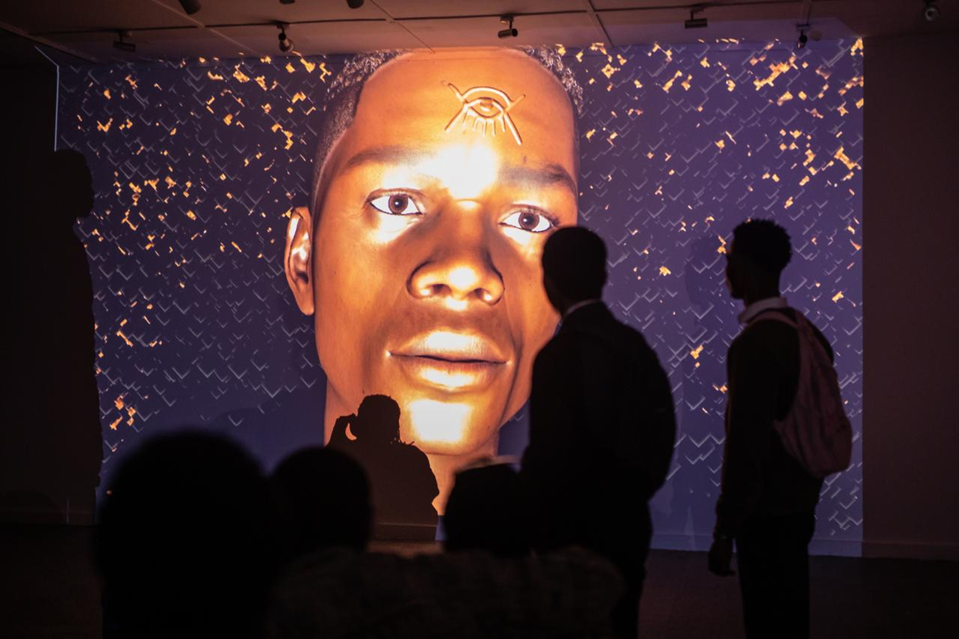 Silhouetted visitors observe a large, golden-toned digital projection of a face with an Eye of Horus symbol.