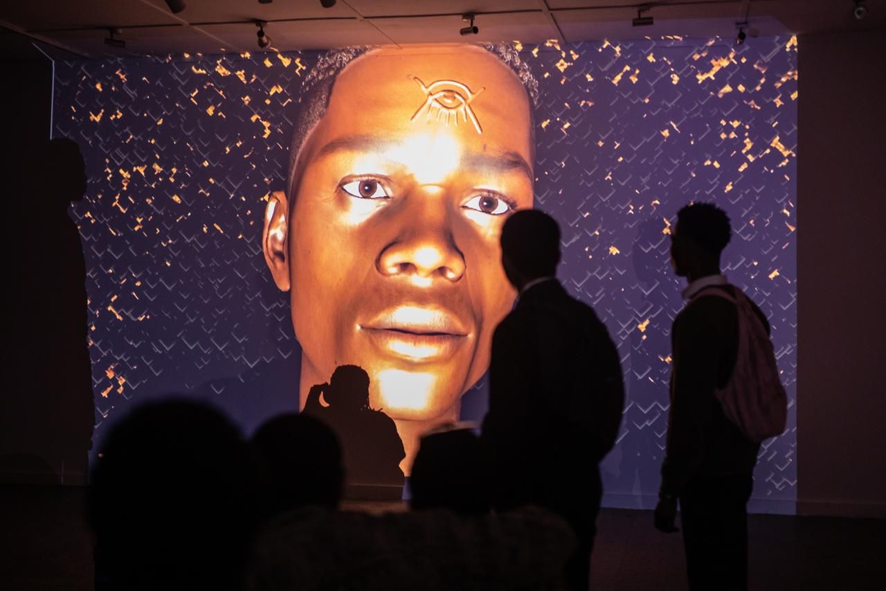 Silhouetted visitors observe a large, golden-toned digital projection of a face with an Eye of Horus symbol.