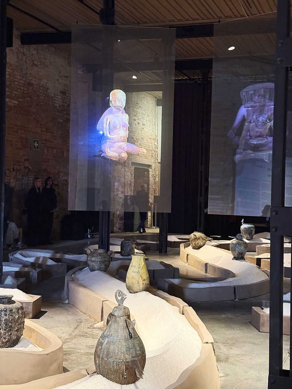 An exhibition with holographic projections of ancient figures above winding paths filled with white granular material and displaying pottery vessels.