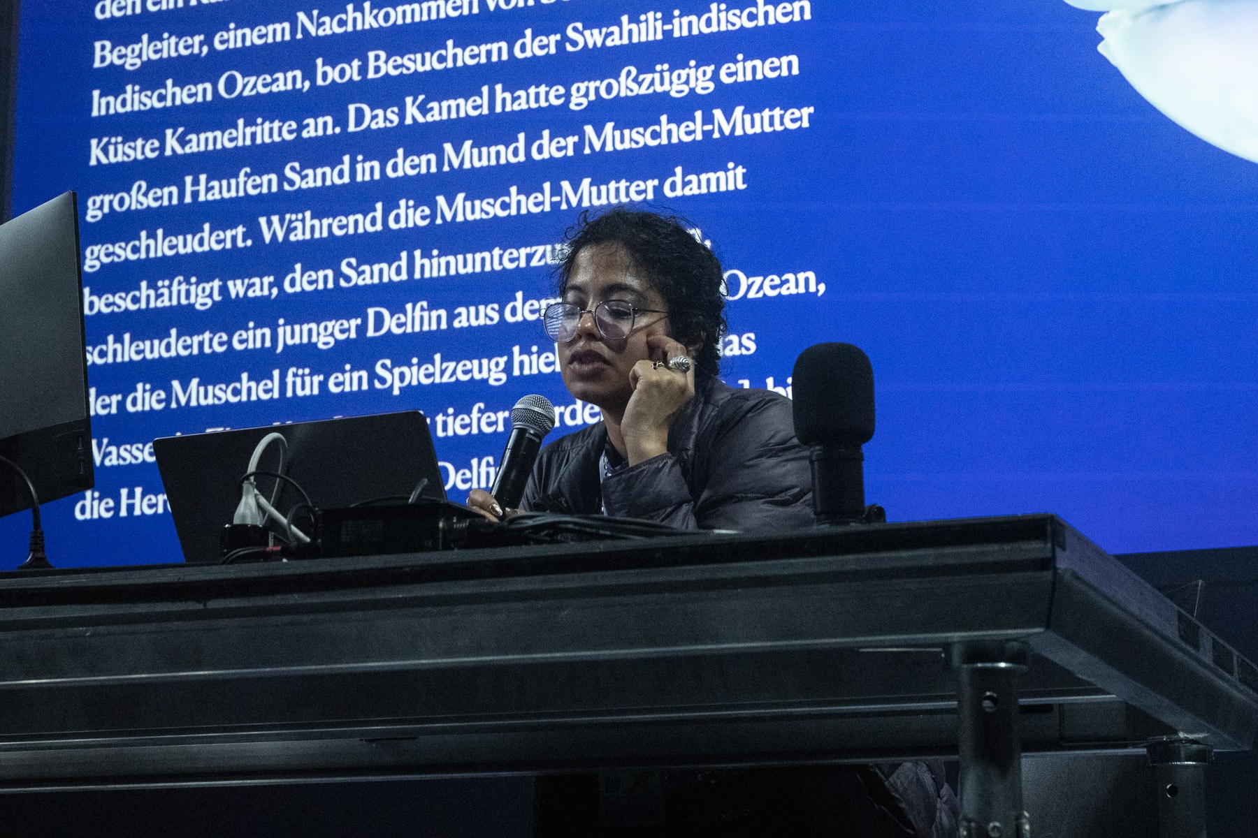 A woman with glasses speaks into a microphone, with a laptop in front and a large blue screen displaying German text behind her.