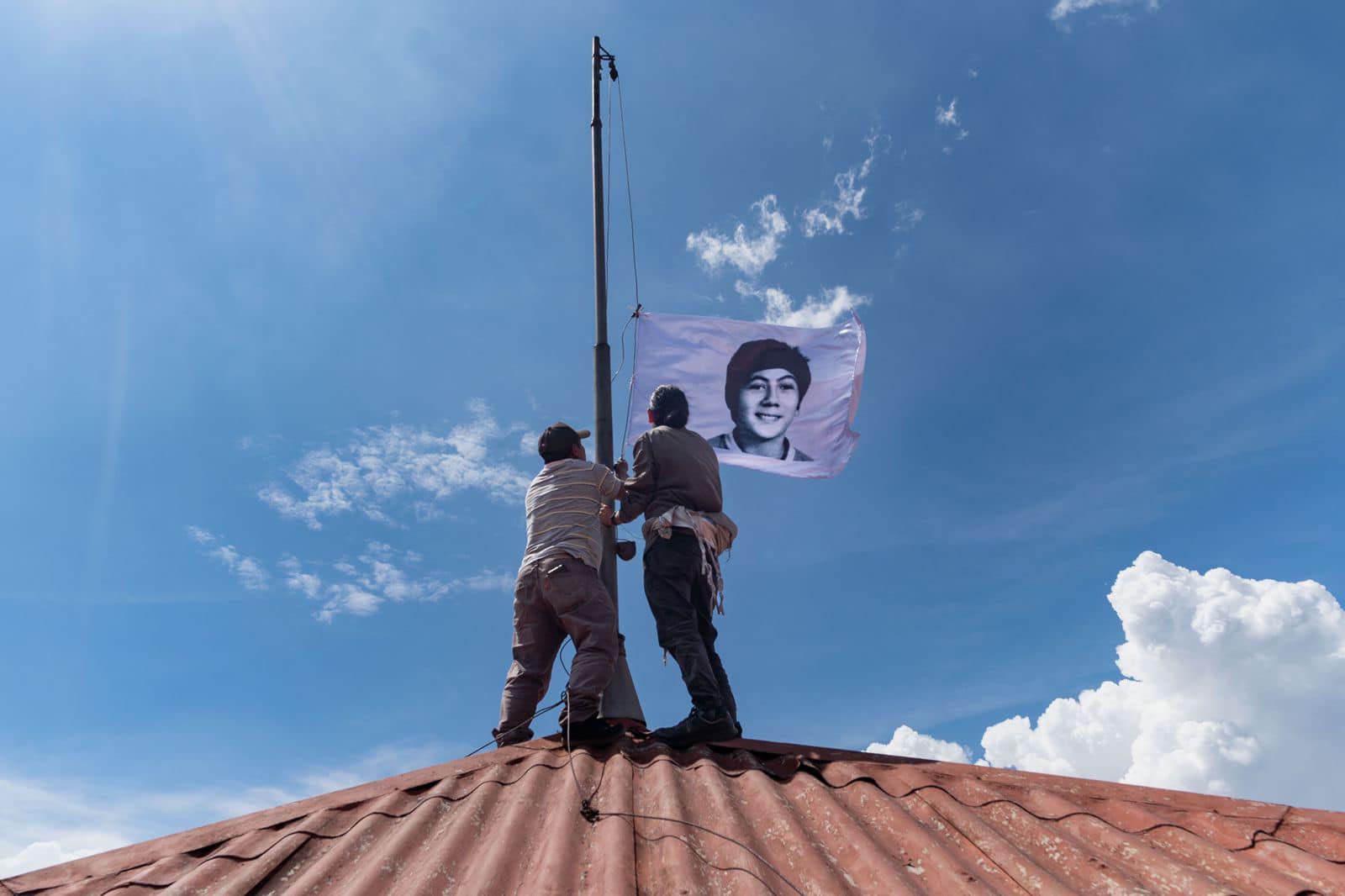 Two people on a tiled roof raise a white flag with a portrait of a smiling young person against a blue sky.