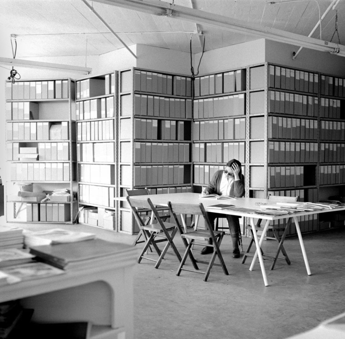 Black and white photo of a person reading at a long table in a room lined with shelves of archival boxes.