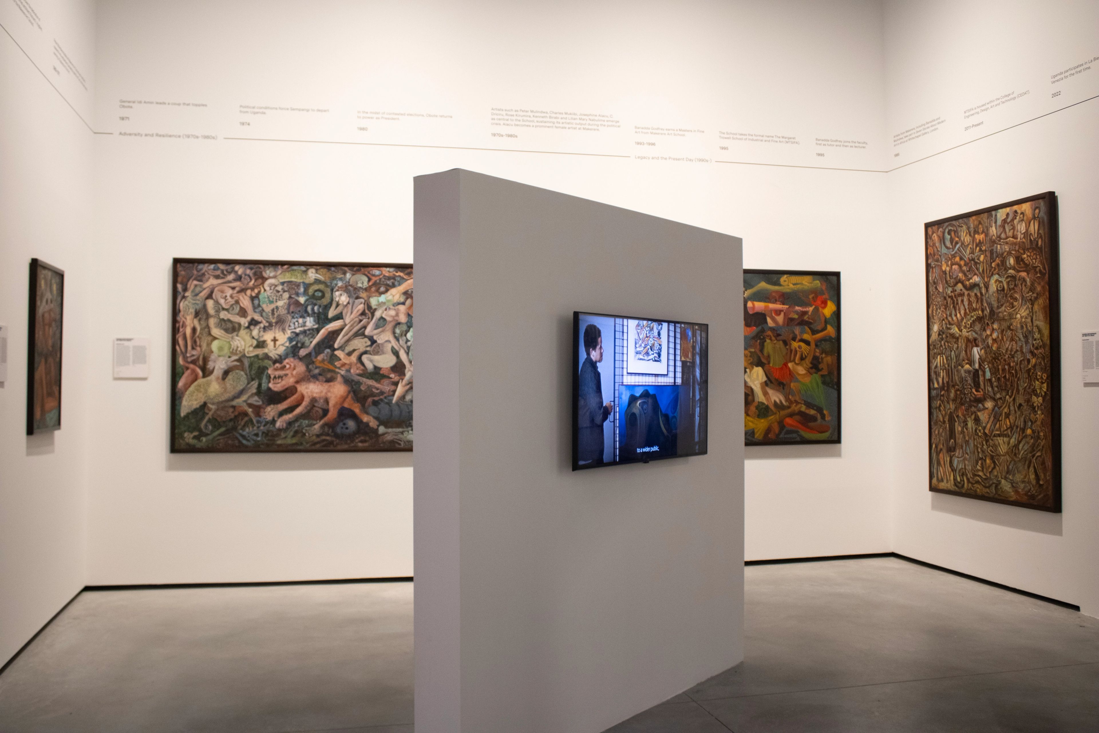 An art gallery with multiple paintings on white walls and a video playing on a screen mounted on a partition.