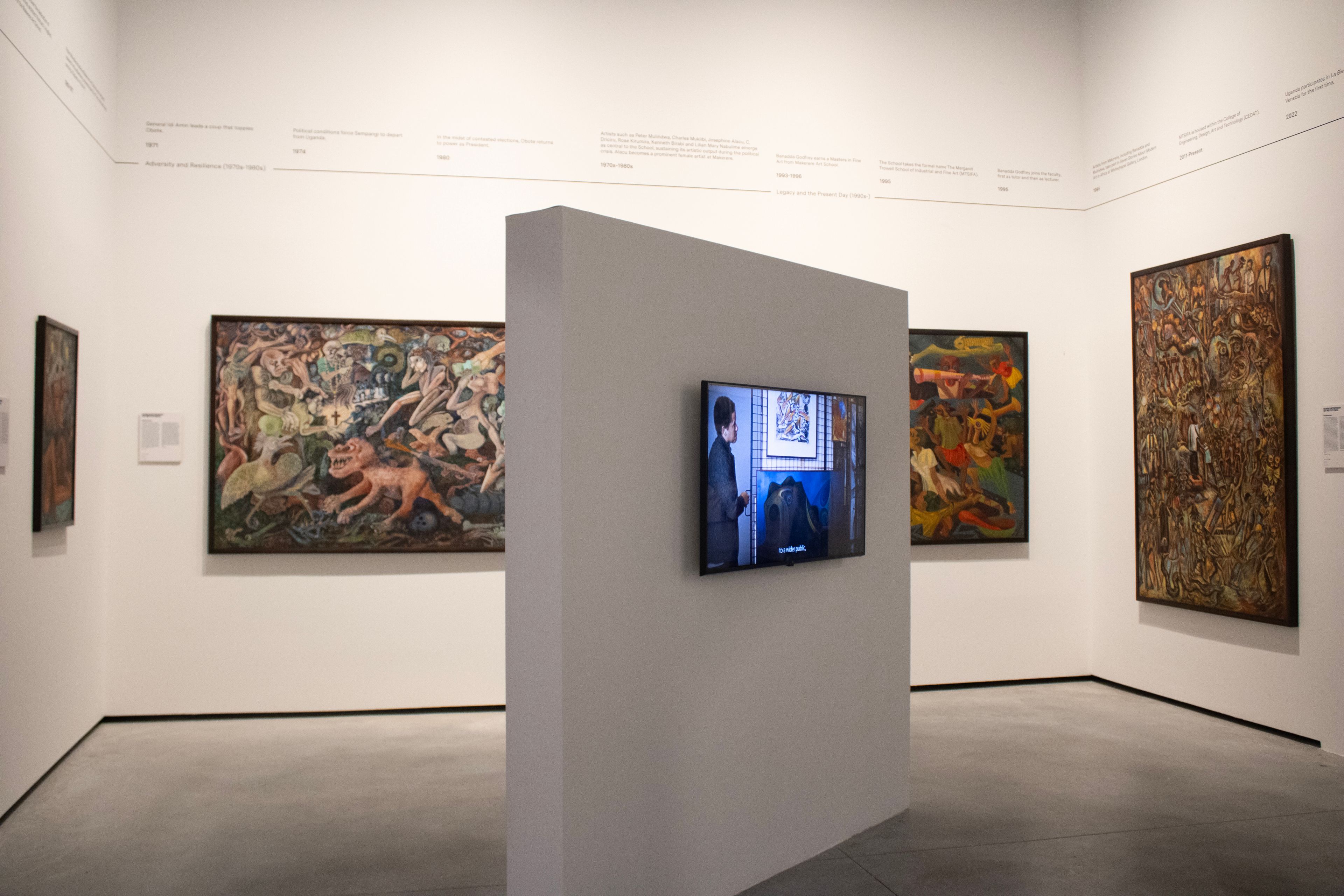An art gallery with multiple paintings on white walls and a video playing on a screen mounted on a partition.