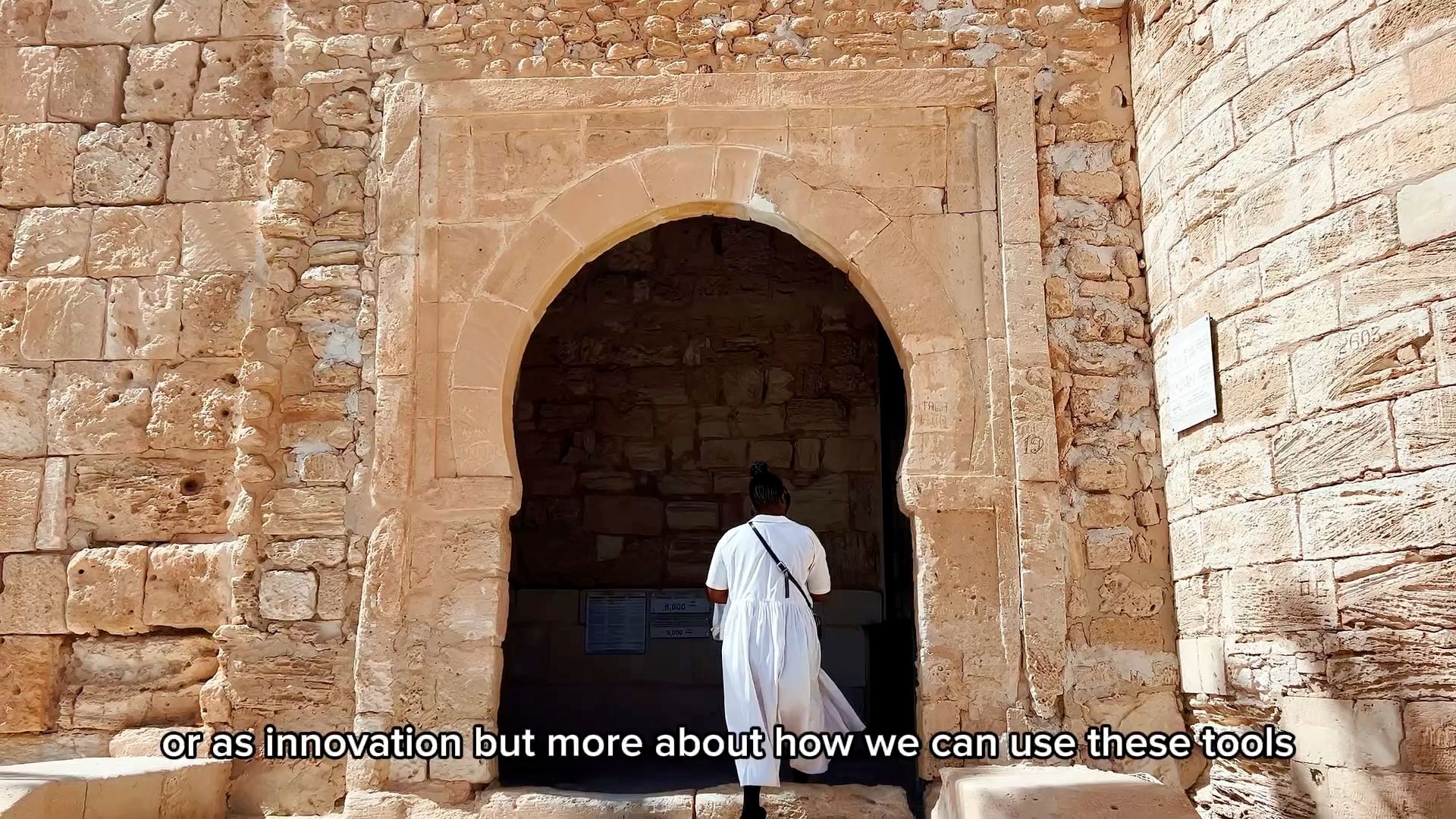A person in a white dress walks into an arched stone doorway.