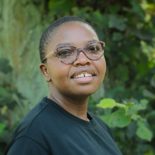 A smiling woman with short dark hair and glasses, wearing a black t-shirt, against a leafy green background.
