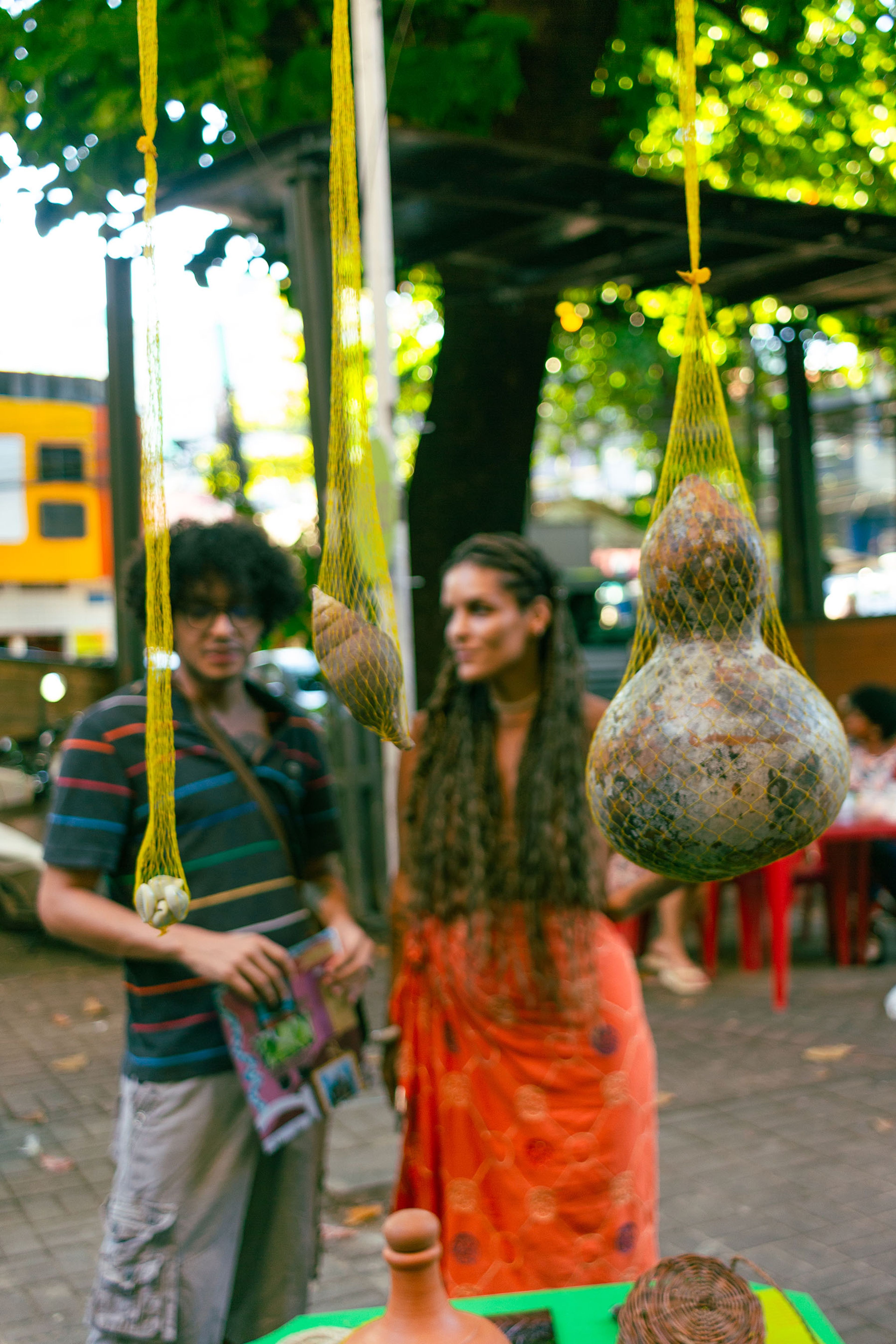 A gourd, a snail shell, and small white shells hang in yellow nets, with two blurred people in the background.