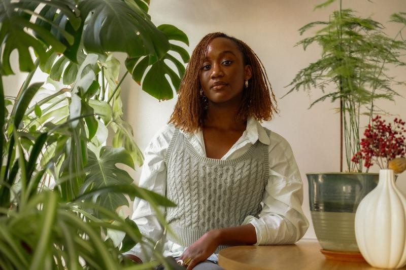 A Black woman with reddish-brown curly hair, wearing a white shirt and grey sweater vest, sits at a table surrounded by green plants.