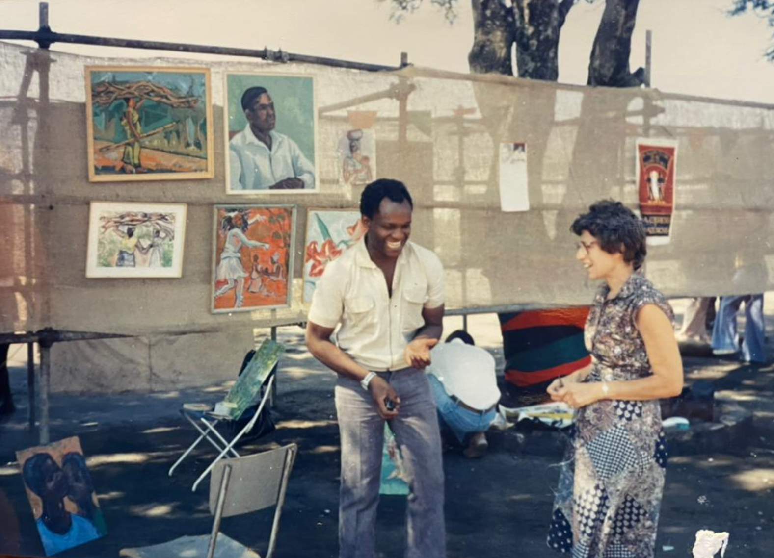 A laughing Black man in a light shirt talks with a smiling White woman at an outdoor art market, with colorful paintings displayed behind them.
