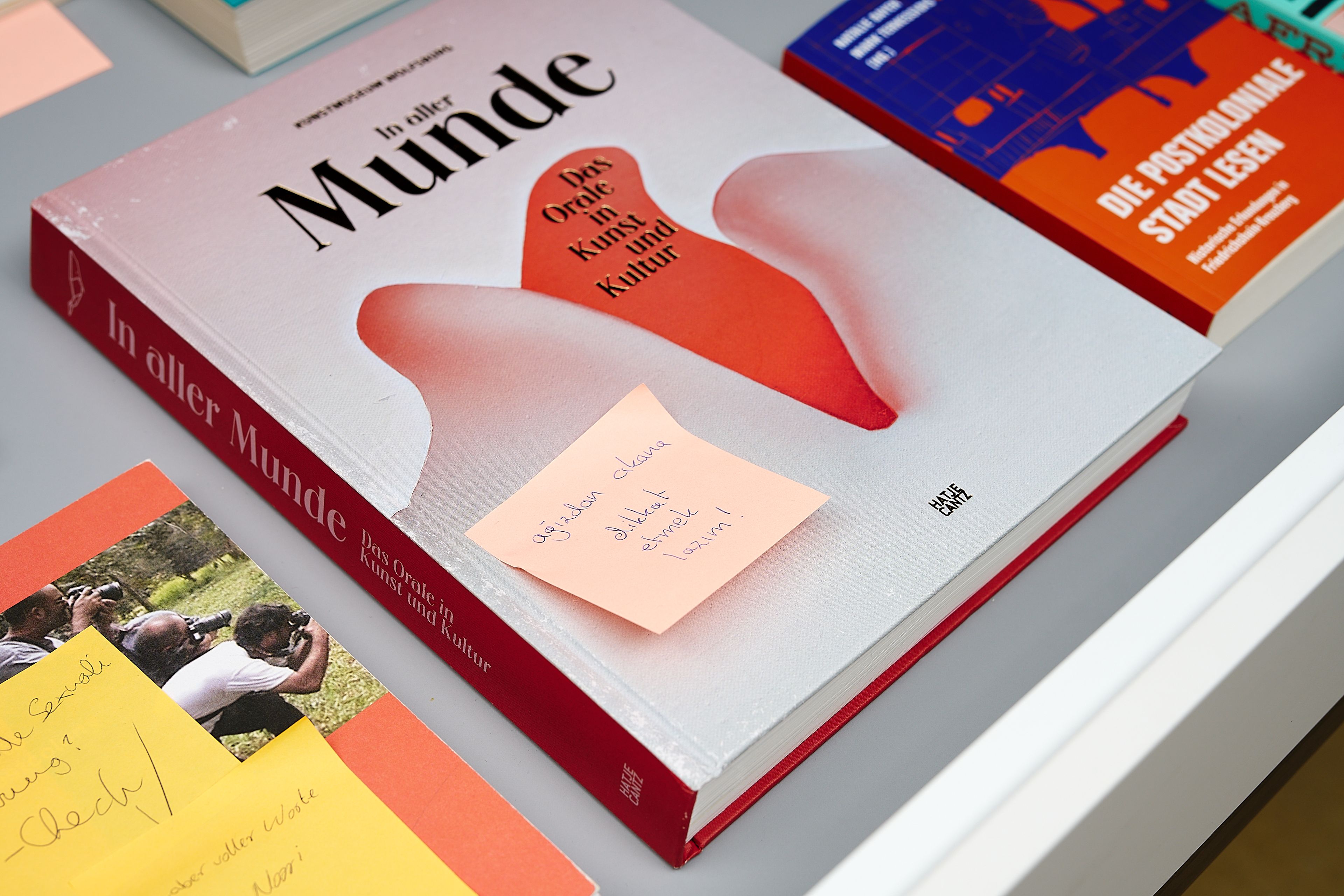 A white book titled "In aller Munde" with red abstract shapes on the cover, featuring a pink sticky note with handwritten text.