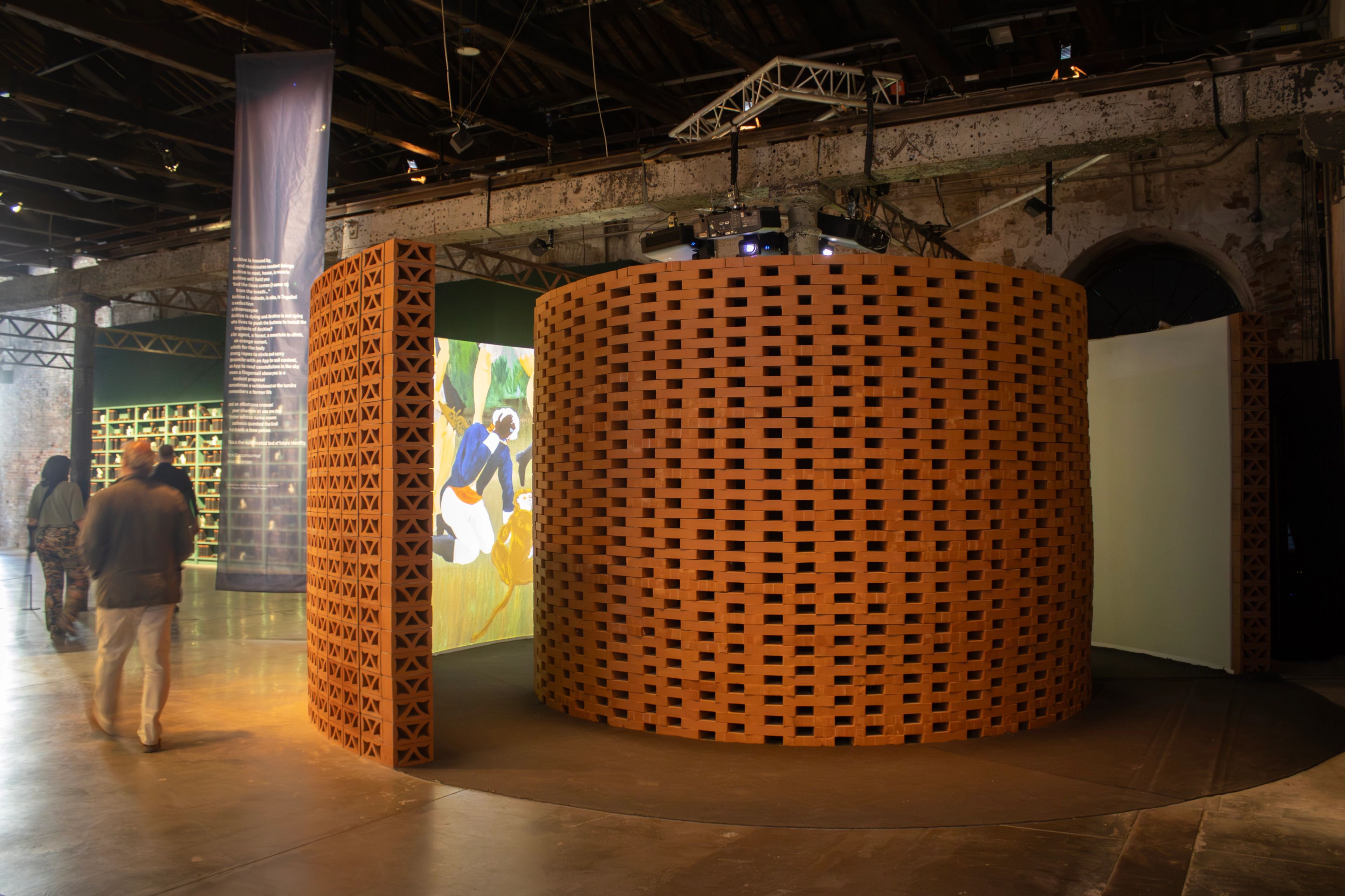 A curved wall of terracotta blocks encloses a large projected artwork in an exhibition space, with visitors nearby.