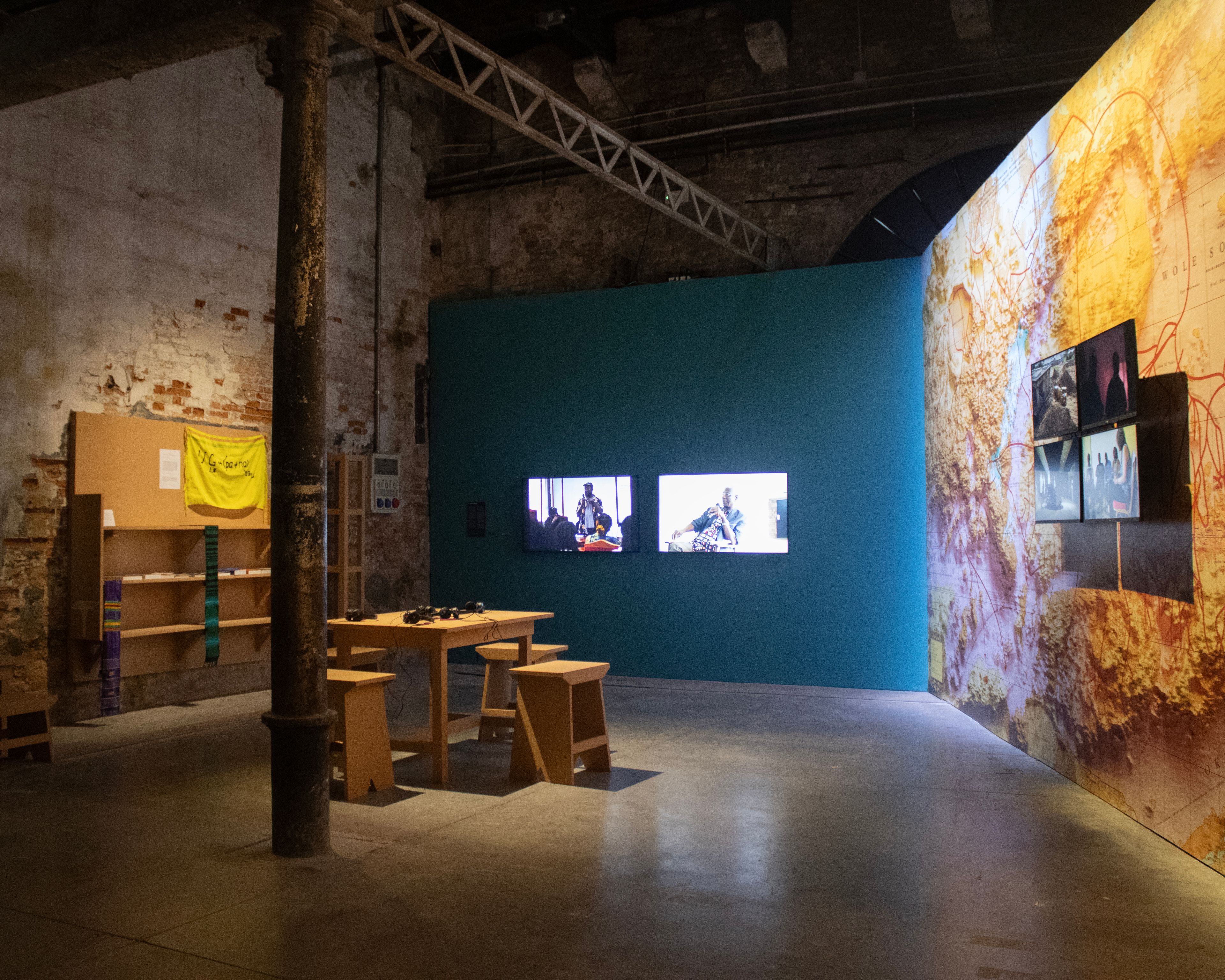Exhibition space with a wooden table, headphones, two video screens on a teal wall, and a large abstract map projection on a right wall with six smaller image displays.
