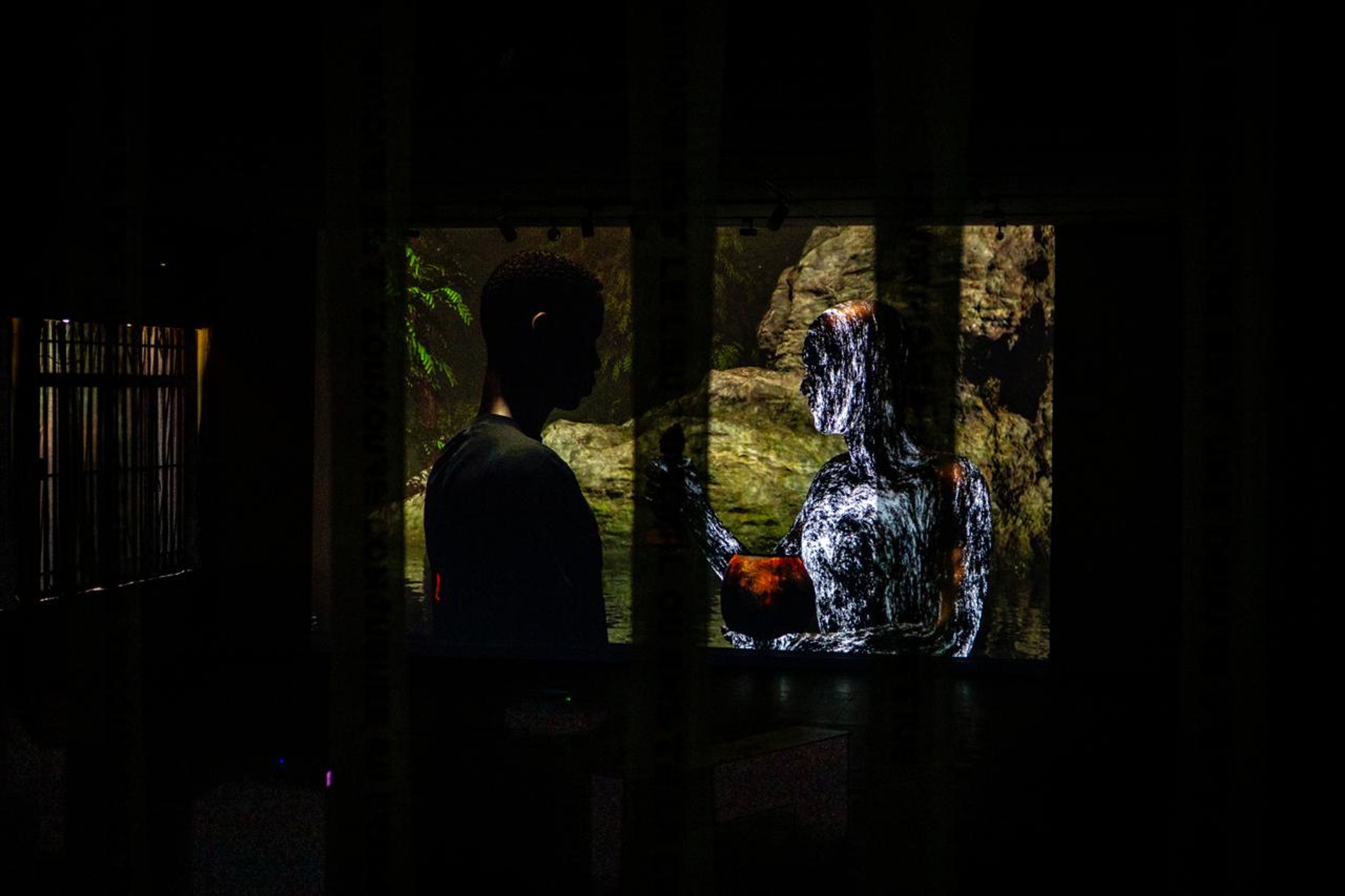 A dark room with a projection showing a silhouetted person facing a textured figure against a rocky backdrop, partially obscured by vertical bars.