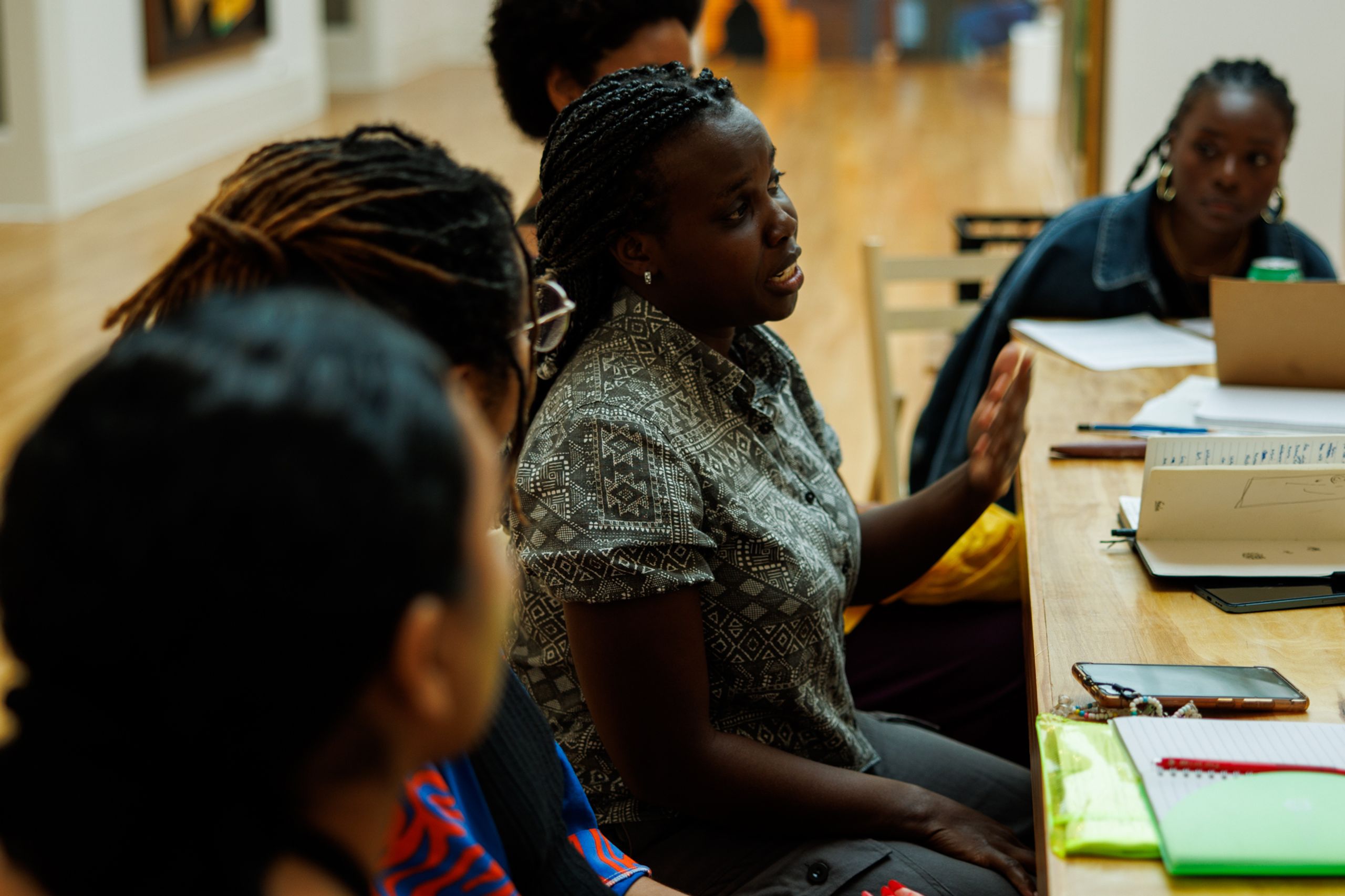 A Black woman speaks and gestures with her hand at a wooden table during a group discussion.