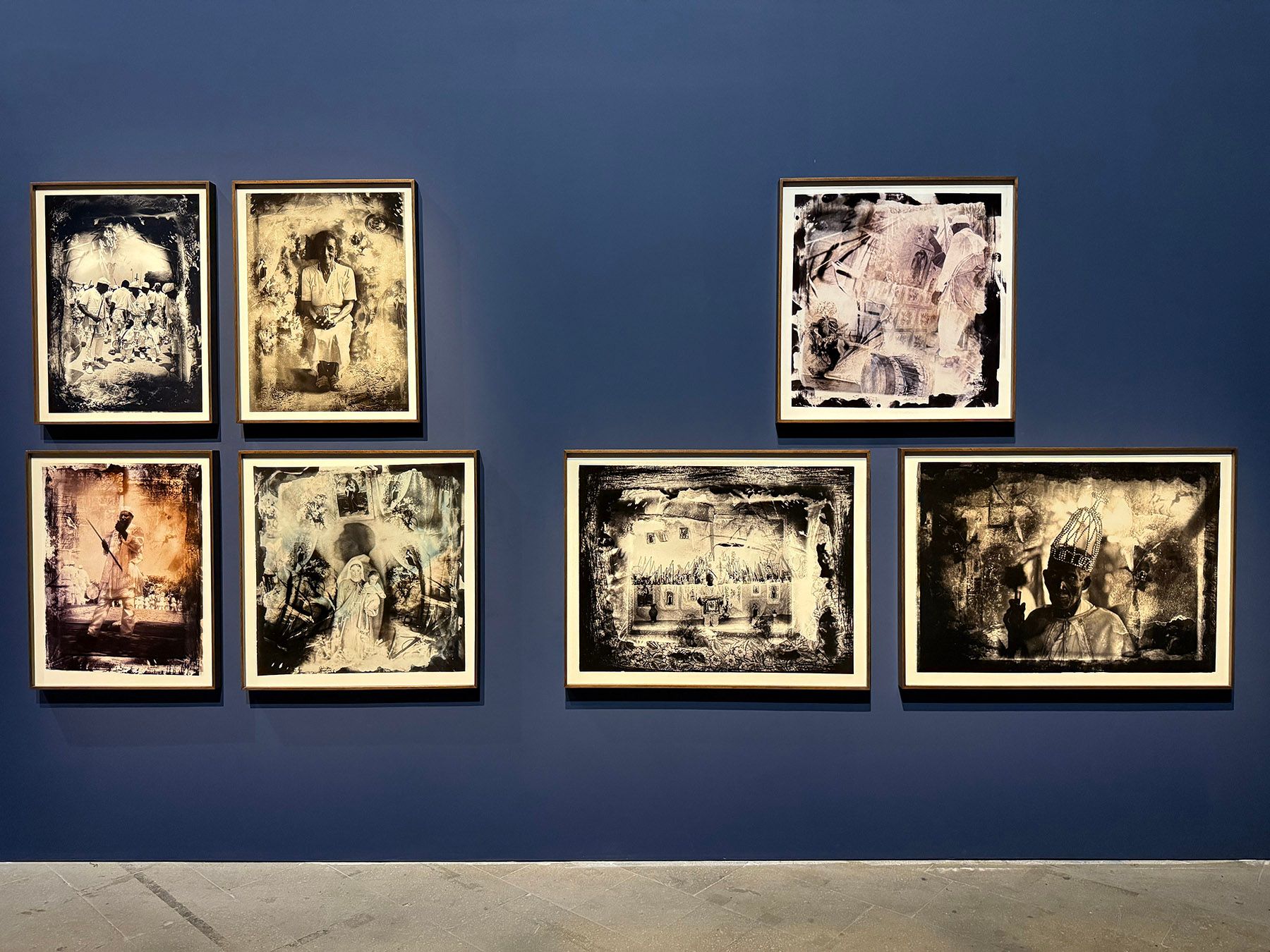 Seven framed, monochrome and sepia-toned artworks with rough white borders, depicting varied subjects, on a dark blue wall.