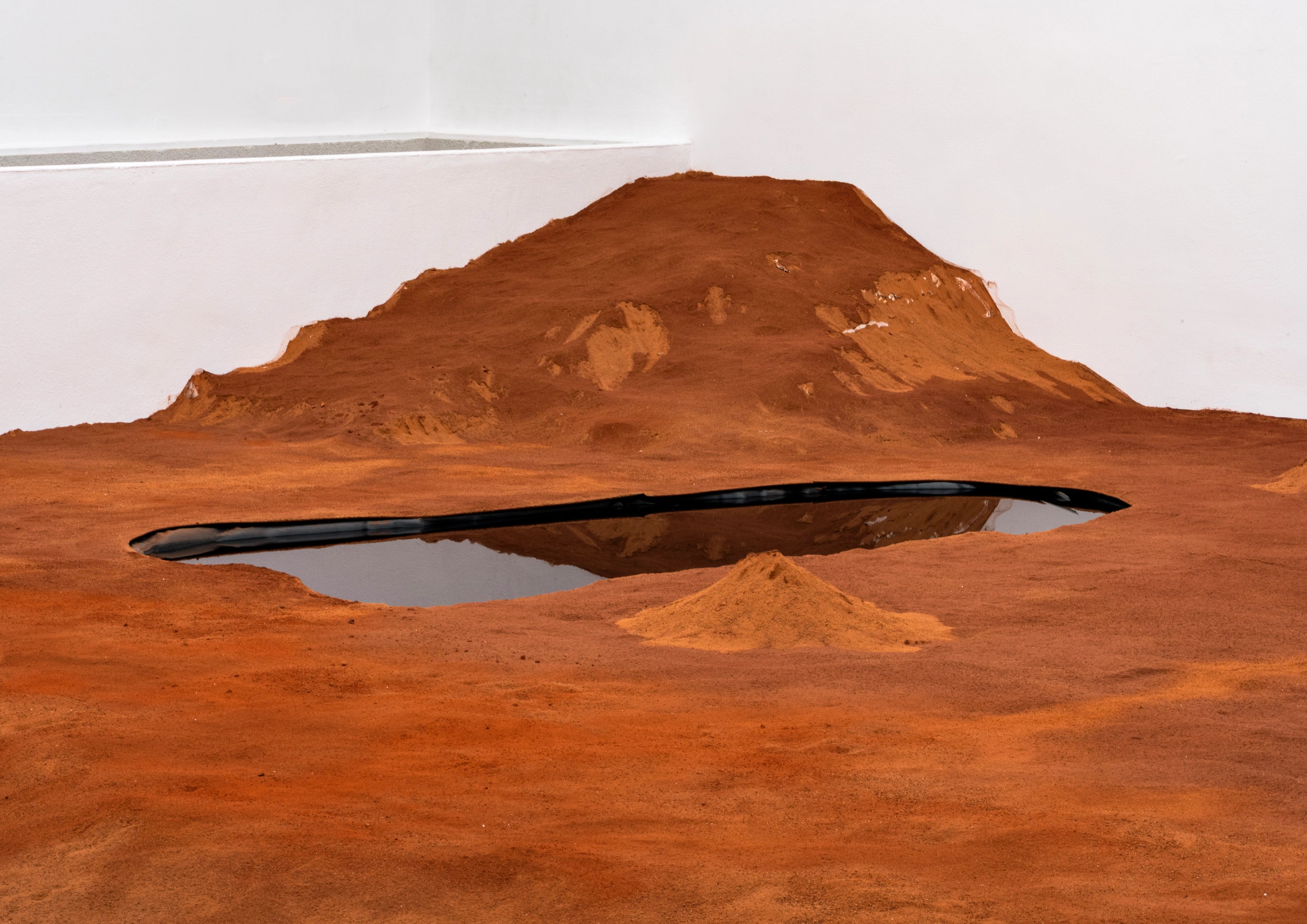 Art installation of reddish-brown sandy hills and a long, dark reflective pool.
