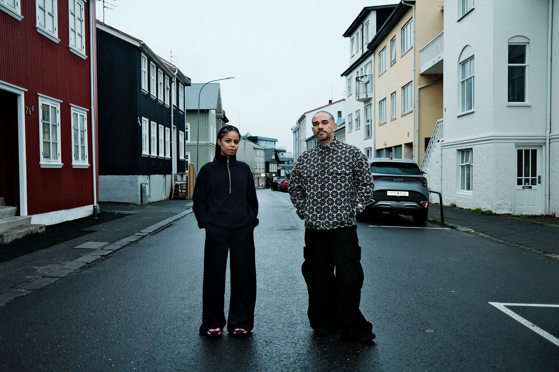 A woman in a black quarter-zip top and wide-leg pants, and a man in a black and white patterned shirt and wide-leg pants, stand on a wet street lined with colorful buildings.