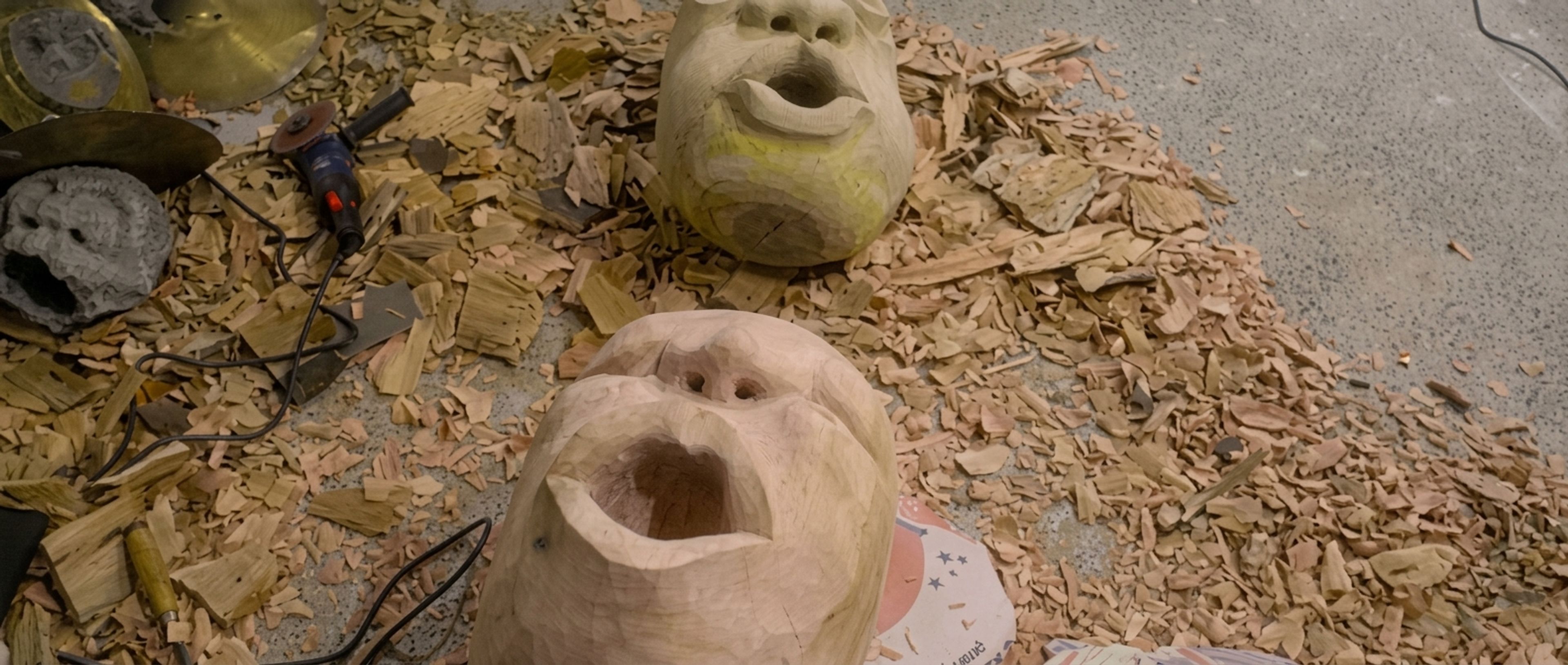 Two unfinished wooden masks with open mouths lie on a floor covered in wood chips, with carving tools visible.