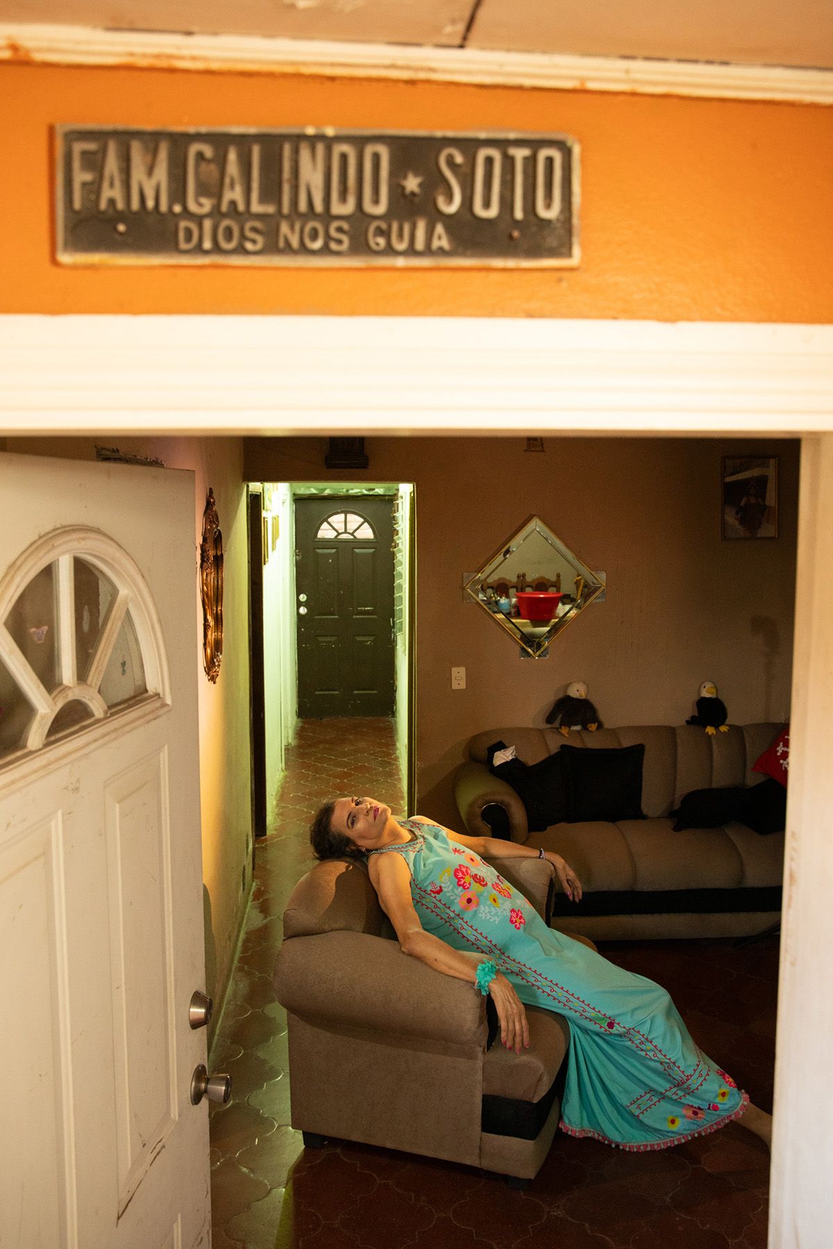 A woman in a blue floral dress reclines on a sofa inside a home, framed by an open doorway with a sign above reading "FAM. GALINDO SOTO DIOS NOS GUIA".
