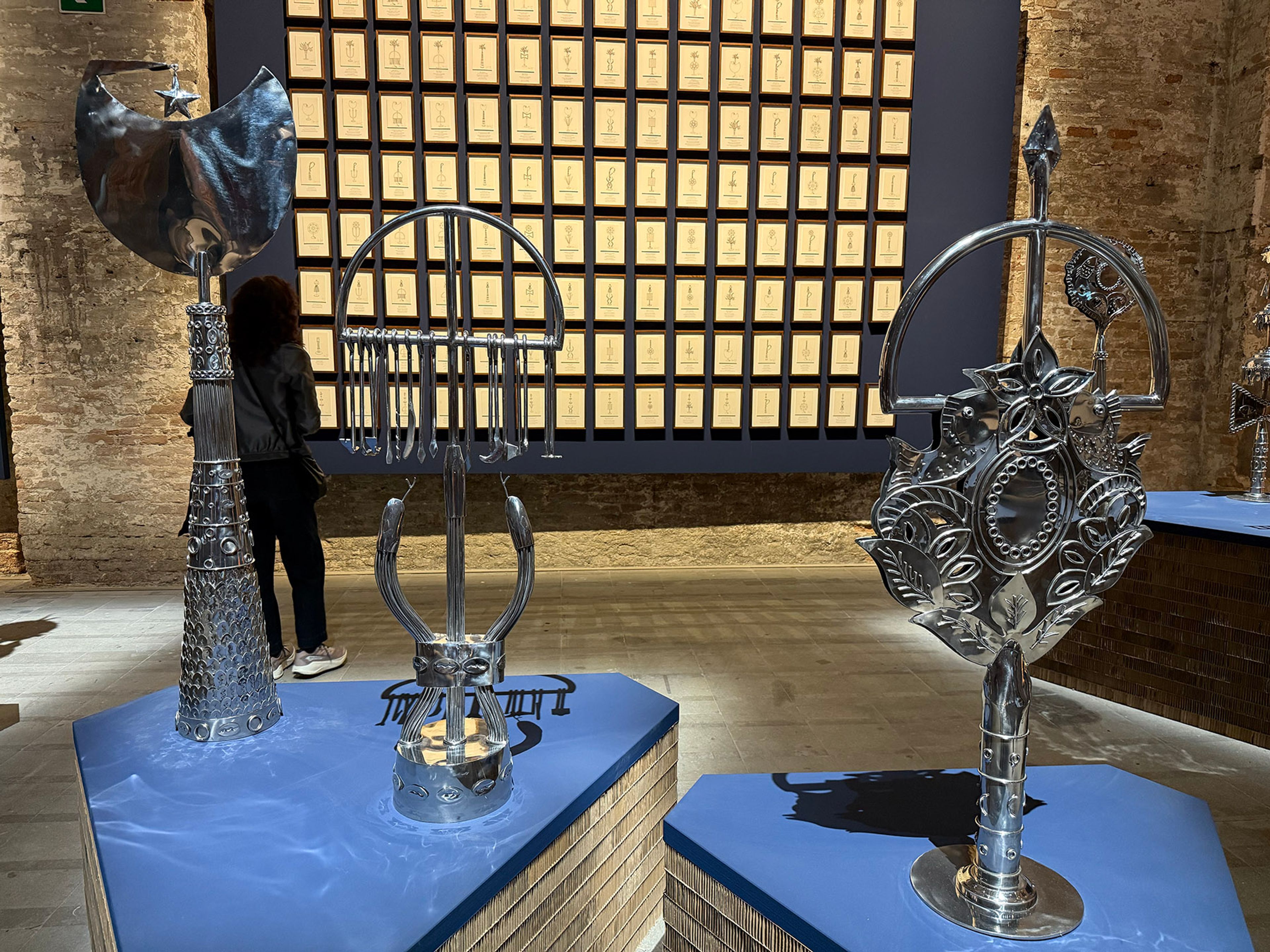 Three ornate metallic sculptures on blue stands in a gallery, with a person viewing them and a grid of framed art on a dark wall.