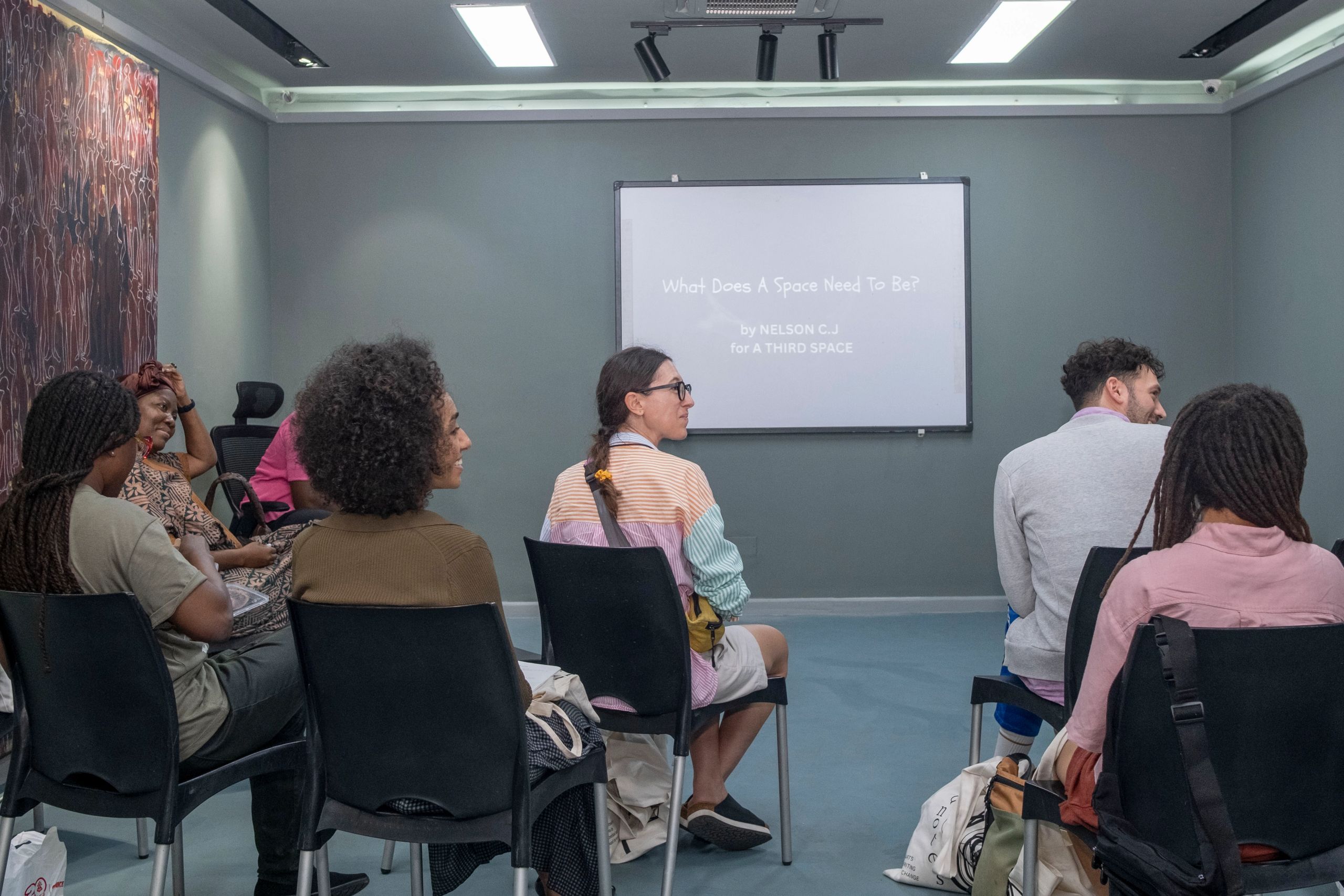 A diverse group of people sits in a modern room, facing a projector screen displaying "What Does A Space Need To Be?".