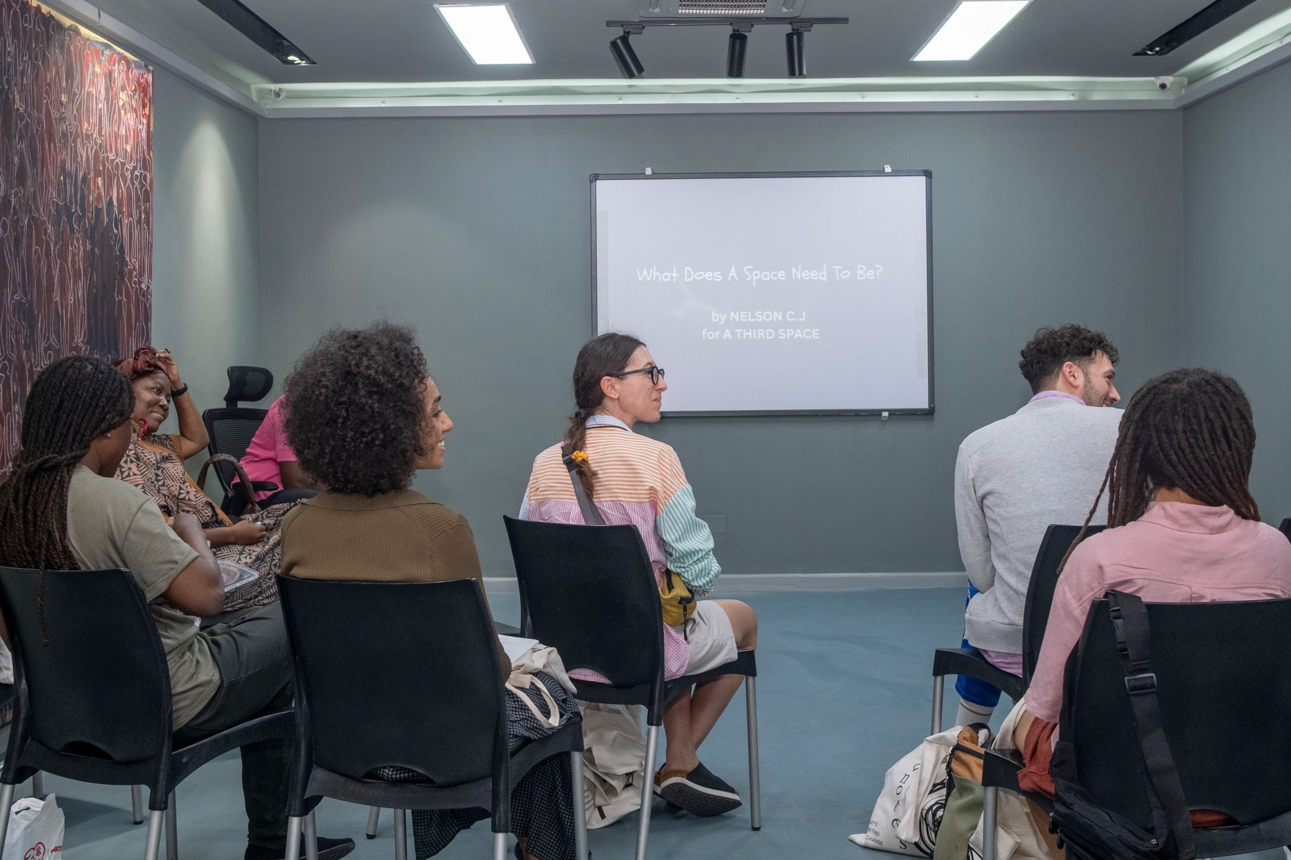 A diverse group of people sits in a modern room, facing a projector screen displaying "What Does A Space Need To Be?".