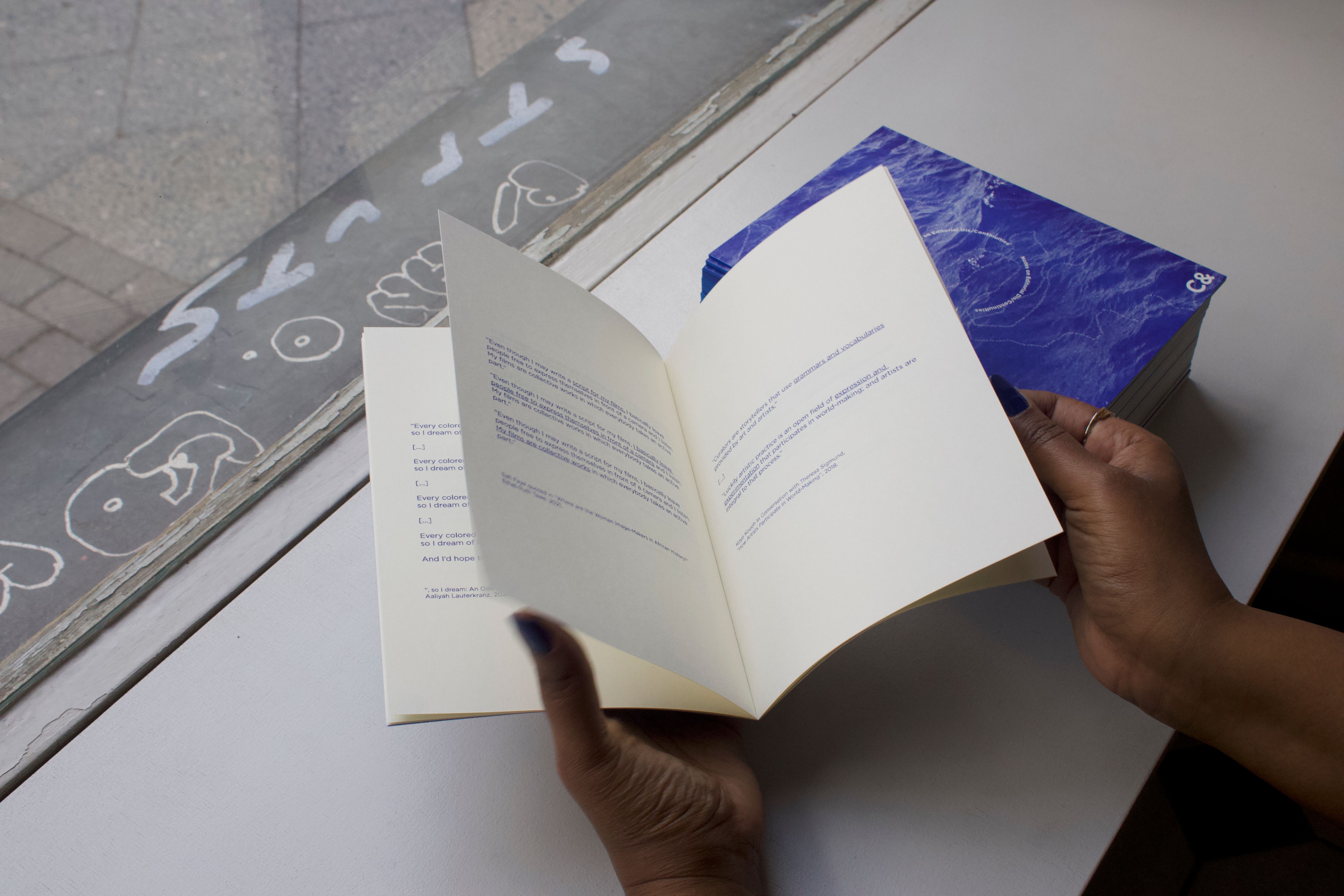 Hands holding an open publication with text, next to a stack of blue books, with graffiti visible through a window in the background.